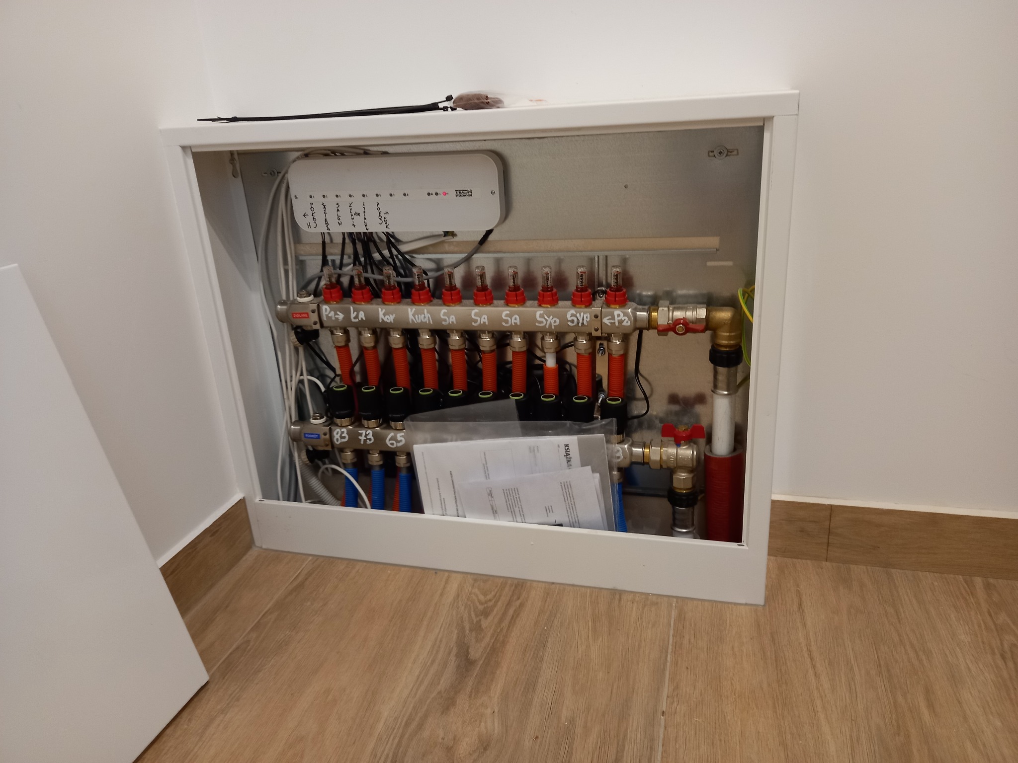 Rozdzielacz ogrzewania podłogowego w białej szafce, z widocznymi rurami, zaworami, sterownikiem TECH i dokumentacją techniczną, zainstalowany przy podłodze wykończonej panelami imitującymi drewno.