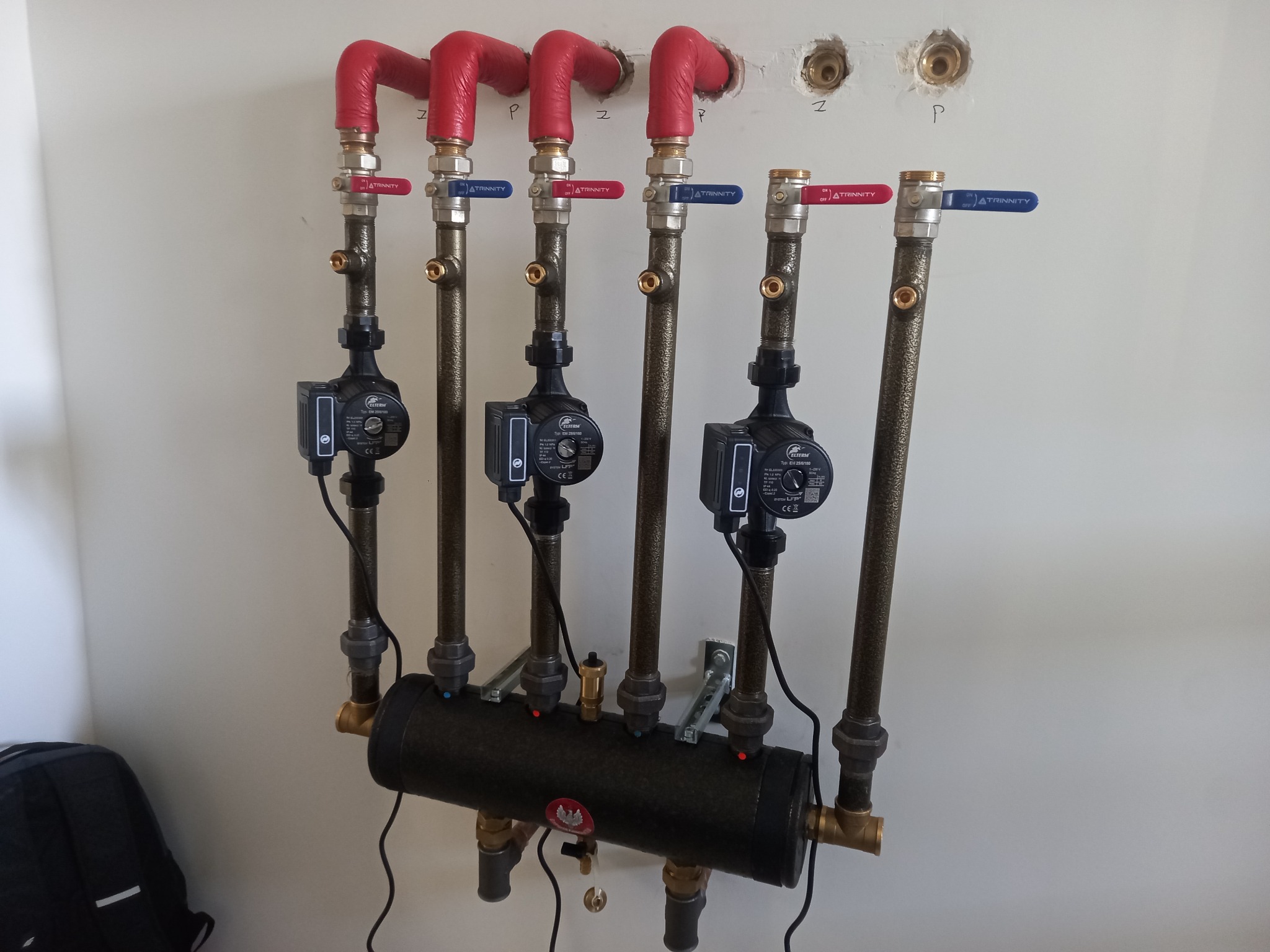 Rozdzielacz hydrauliczny z pompami obiegowymi, rury miedziane z czerwonymi izolacjami na kolankach, zawory kulowe z czerwonymi i niebieskimi uchwytami, montaż na białej ścianie z widocznymi...