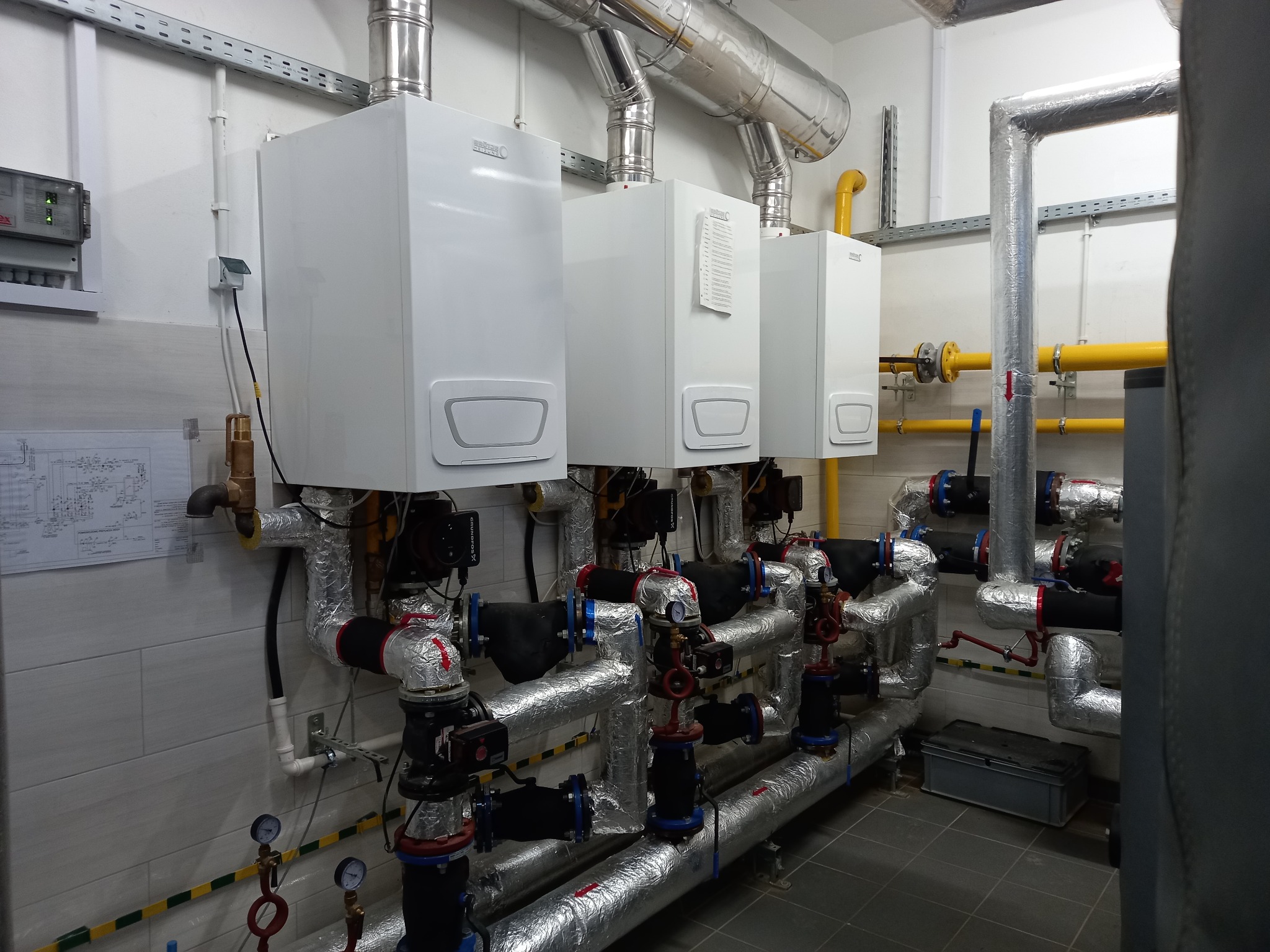 Trzy identyczne, białe piece gazowe zamontowane na ścianie z szarą glazurą, z widocznymi rurami i zaworami, izolowanymi folią aluminiową, w pomieszczeniu technicznym.