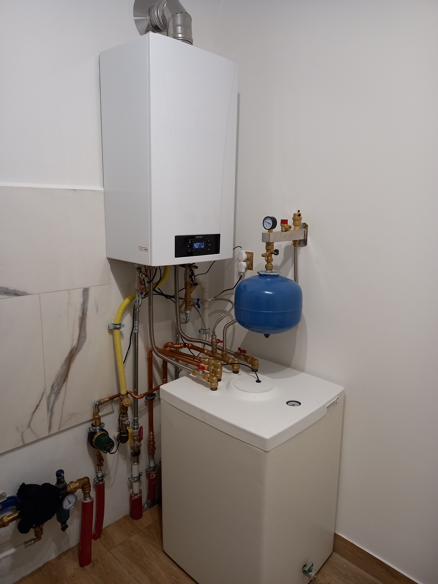 Nowoczesny, biały piec gazowy kondensacyjny zamontowany w rogu pomieszczenia z widocznymi rurami miedzianymi, żółtym przewodem gazowym i niebieskim zbiornikiem wyrównawczym.