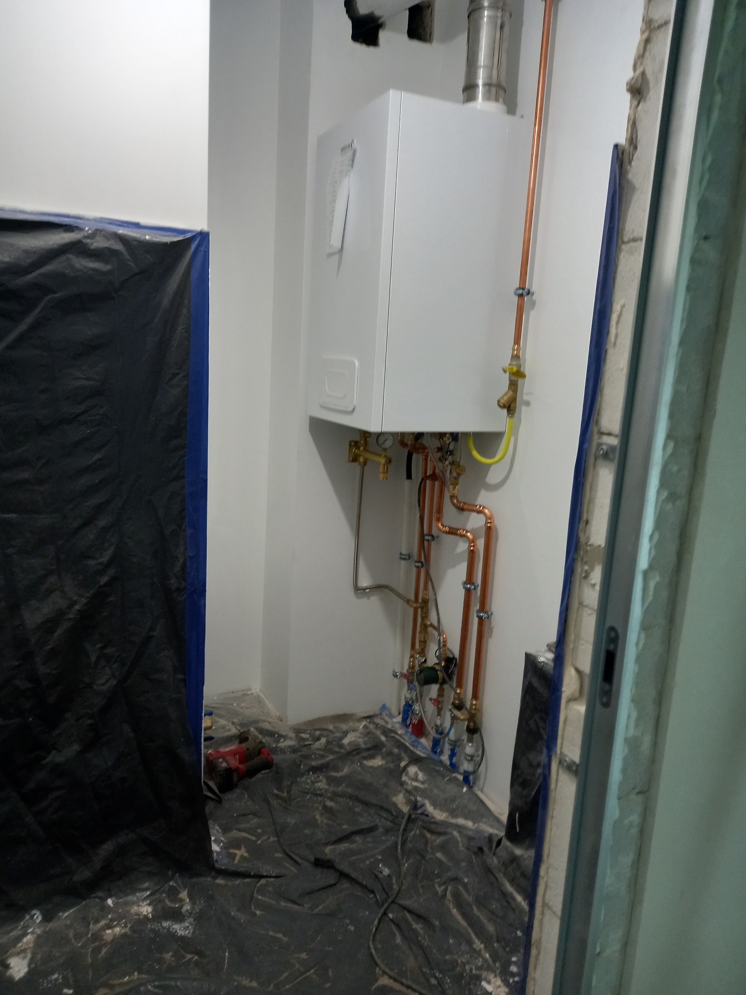 Biały piec gazowy zamontowany na ścianie, z widocznymi miedzianymi rurami i zaworami, zabezpieczone podłoże czarną folią malarską, stan surowy pomieszczenia.