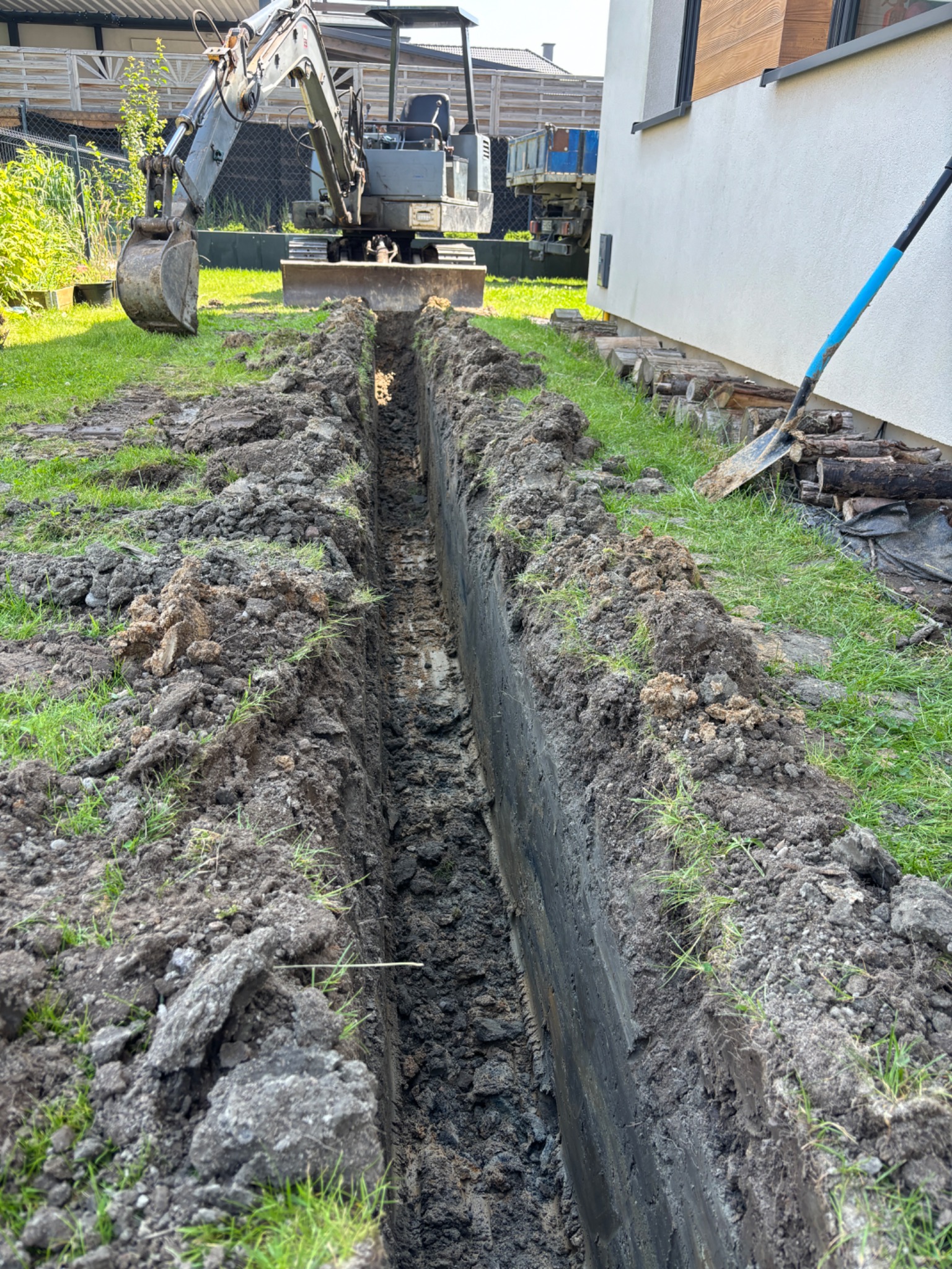 Wykop wąskoprzestrzenny wykonany koparką obok budynku, z widoczną łopatą i stertą drewna, w tle zaparkowany samochód dostawczy.