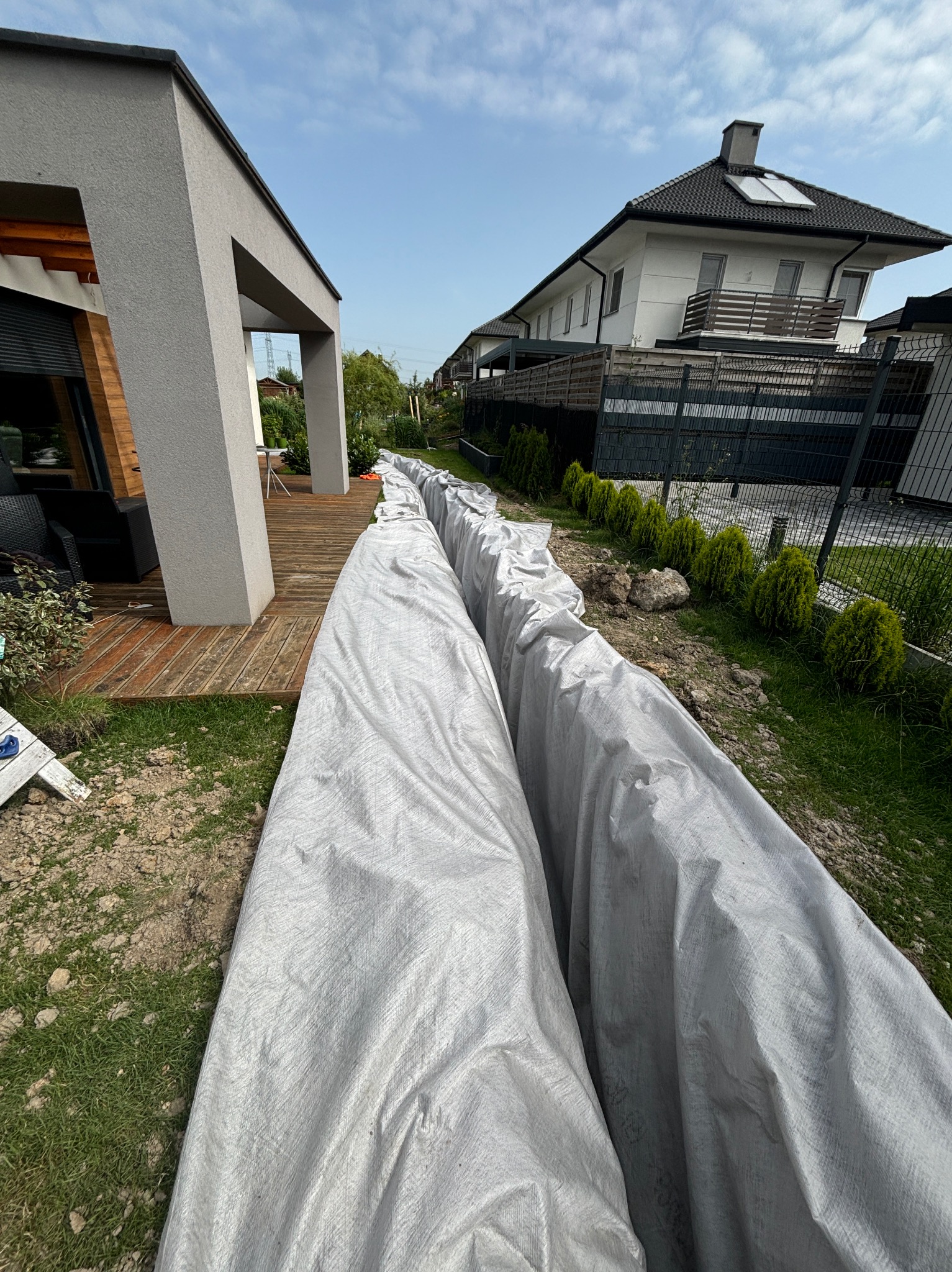 Długi wykop pod fundamenty zabezpieczony szarą folią budowlaną, widok z góry na tle nowoczesnego domu z drewnianym tarasem i ogrodem z tujami.