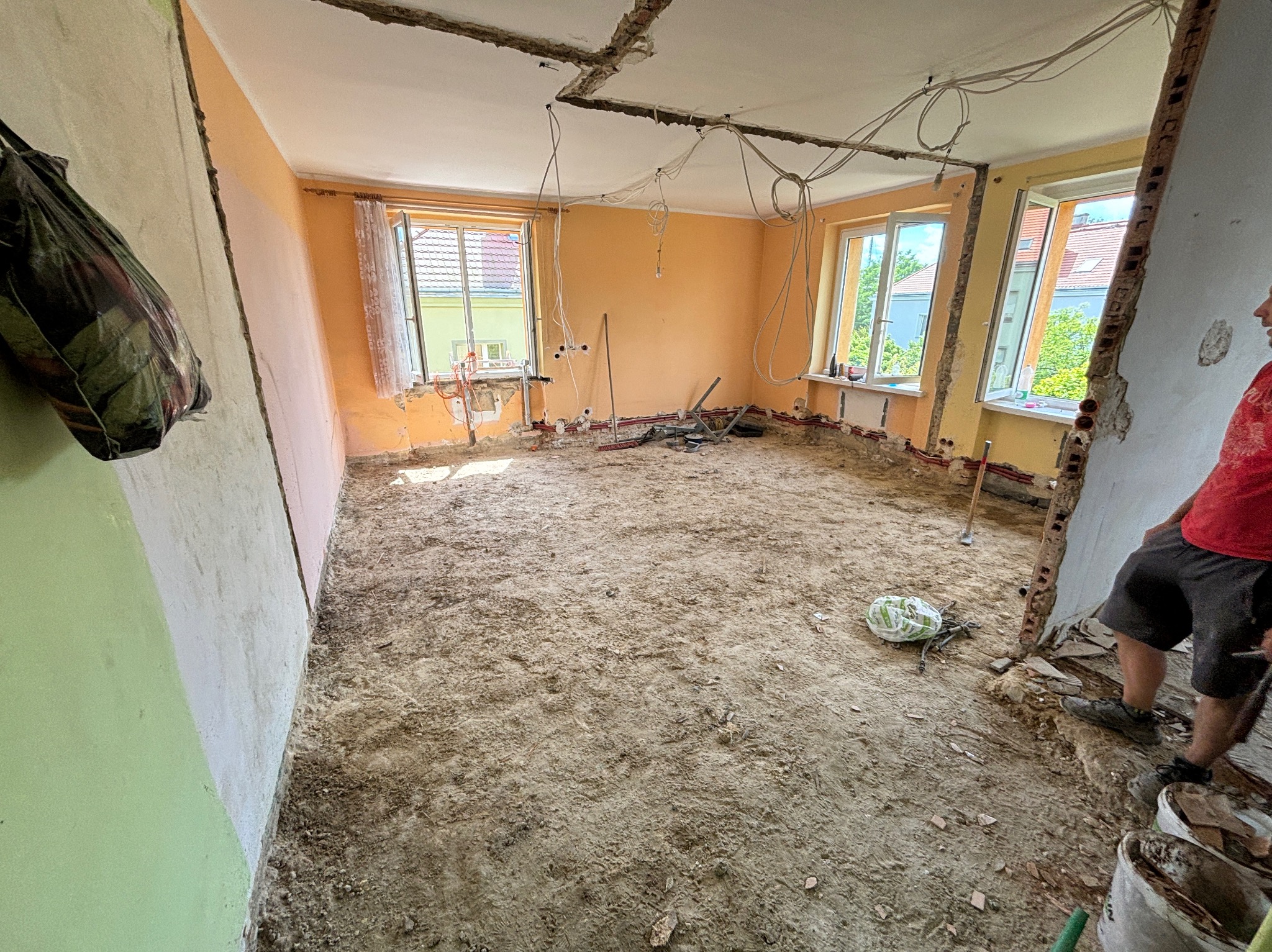 Wnętrze pokoju w trakcie wyburzania ścian, widoczne odsłonięte przewody elektryczne, gruz na podłodze, fragment ściany z resztkami tapety i widok z okna na budynki mieszkalne.