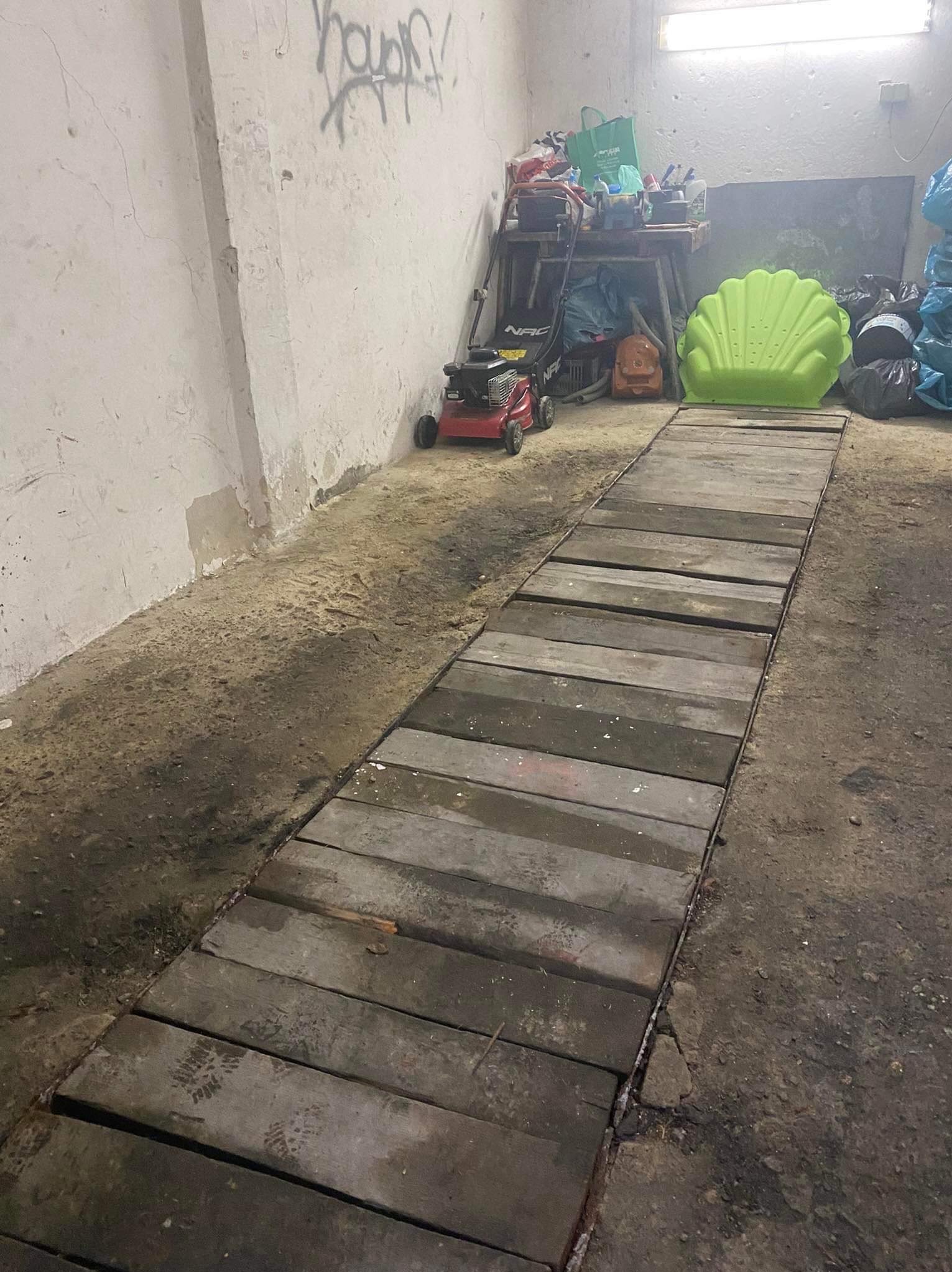 Tymczasowa drewniana ścieżka ułożona na ziemi w pomieszczeniu gospodarczym, w tle regał z narzędziami, zabawkową kosiarką i zieloną muszlą.