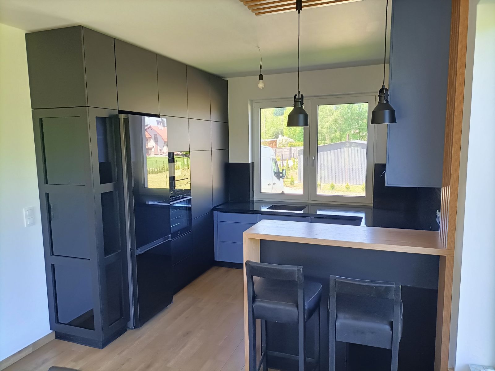 Nowoczesna kuchnia w odcieniach szarości z drewnianym blatem baru i czarnymi hokerami, oświetlona wiszącymi lampami z metalowymi kloszami. Widoczne okno z widokiem na zieleń.