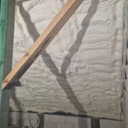 Ściana poddasza pokryta warstwą jasnej pianki izolacyjnej, widoczne elementy konstrukcji drewnianej i przewody elektryczne na podłodze.