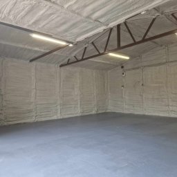 Z.U.A EKTOR - Wnętrze garażu z izolacją natryskową pianką na ścianach i suficie, widoczne metalowe elementy konstrukcyjne dachu i oświetlenie jarzeniowe, szara posadzka betonowa.