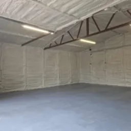 Wnętrze garażu z izolacją natryskową pianką na ścianach i suficie, widoczne metalowe elementy konstrukcyjne dachu i oświetlenie jarzeniowe, szara posadzka betonowa.