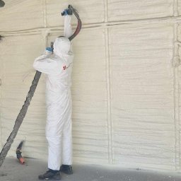 Z.U.A EKTOR - Pracownik w kombinezonie ochronnym natryskuje pianę izolacyjną na ścianę wewnątrz budynku z metalową konstrukcją dachu.