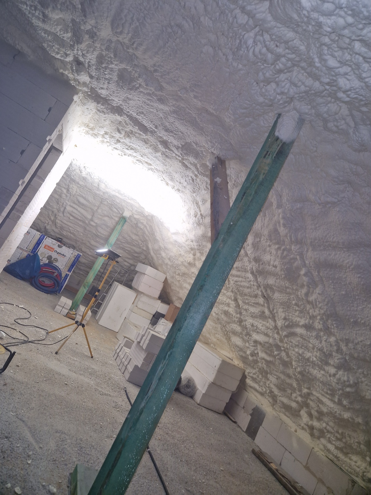 Poddasze z widoczną strukturą natryśniętej pianki izolacyjnej, wspartej metalowym profilem, z widocznym oświetleniem roboczym i materiałami budowlanymi w tle.