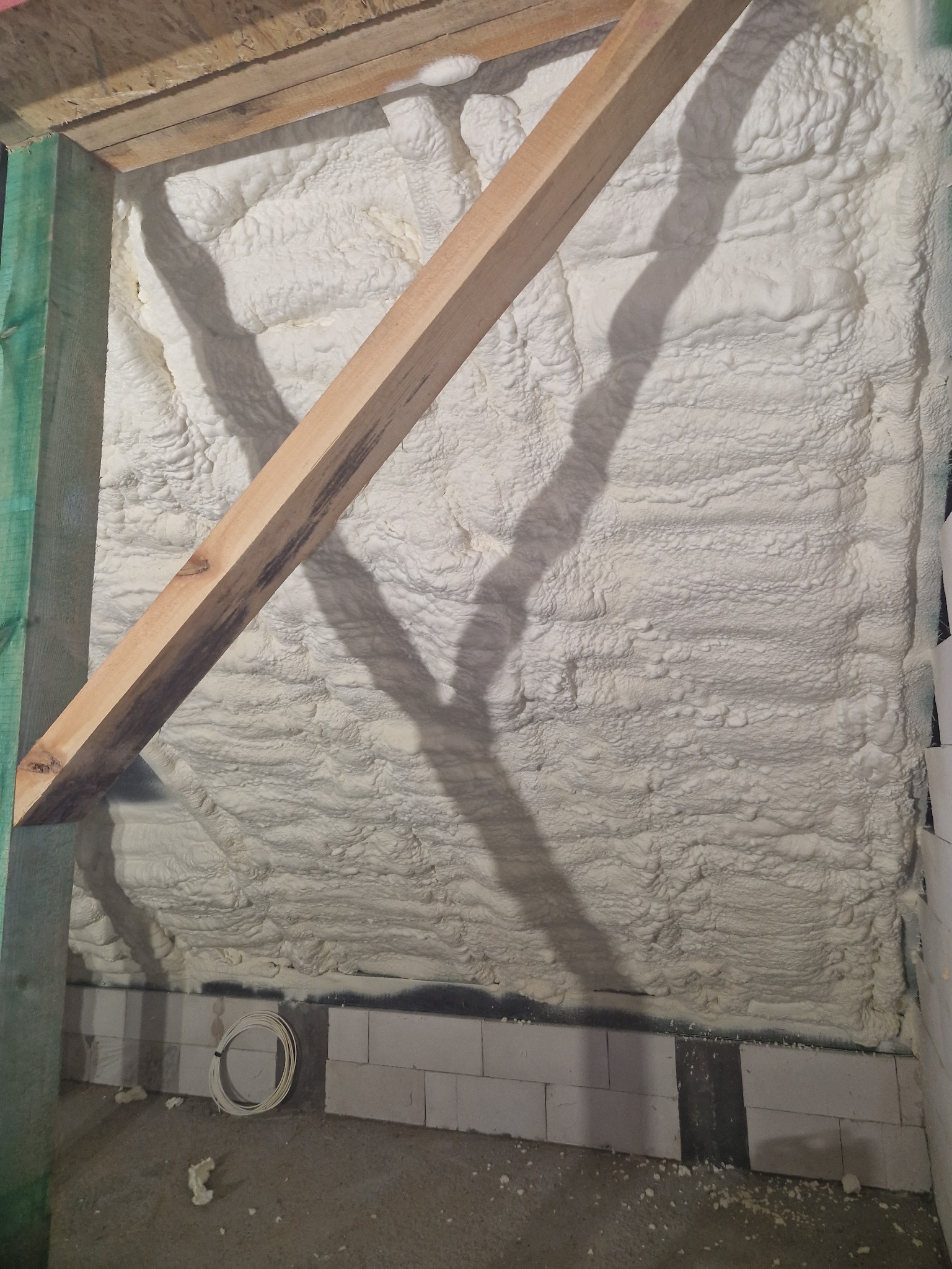 Ściana poddasza pokryta warstwą jasnej pianki izolacyjnej, widoczne elementy konstrukcji drewnianej i przewody elektryczne na podłodze.