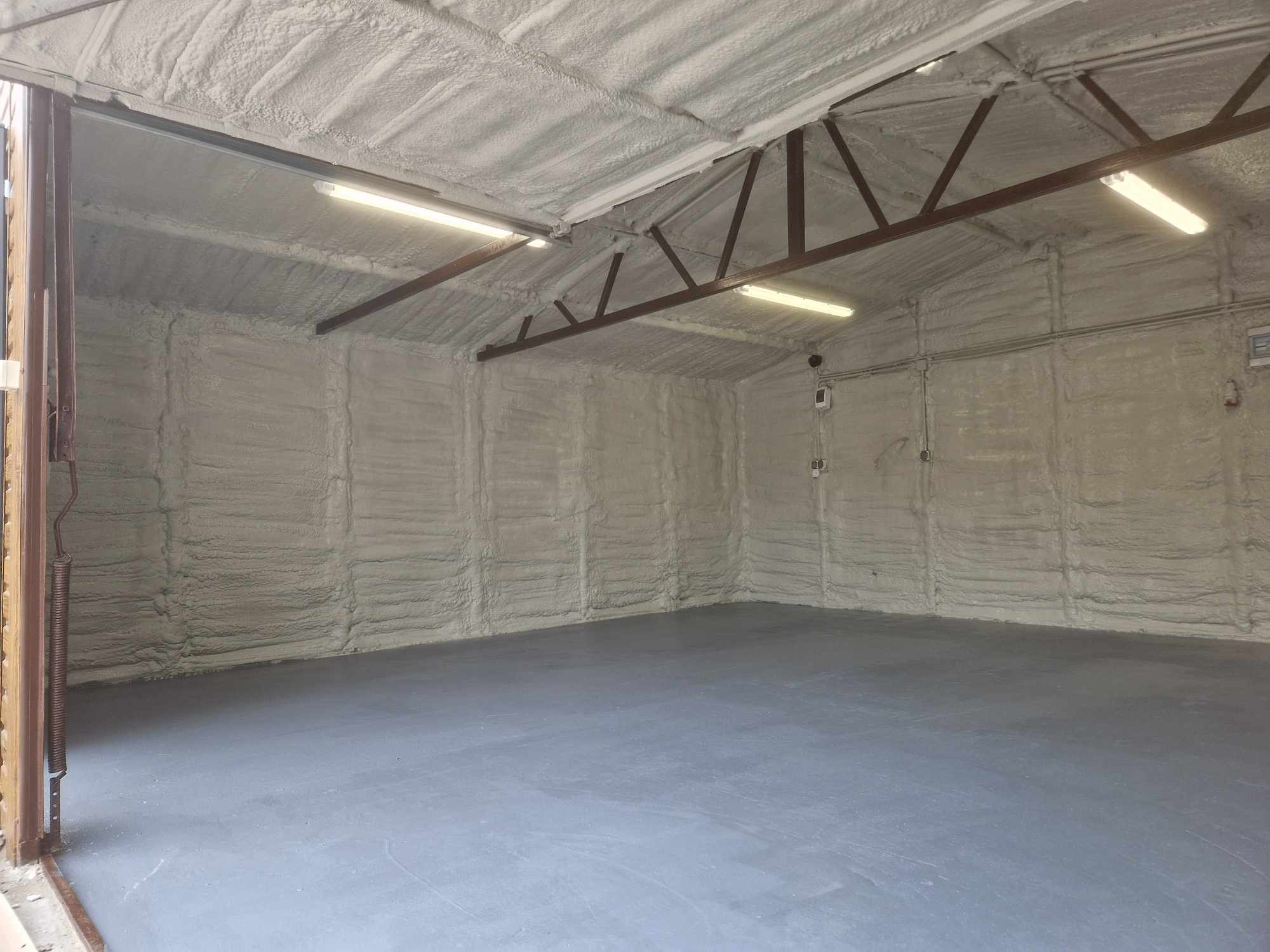 Wnętrze garażu z izolacją natryskową pianką na ścianach i suficie, widoczne metalowe elementy konstrukcyjne dachu i oświetlenie jarzeniowe, szara posadzka betonowa.