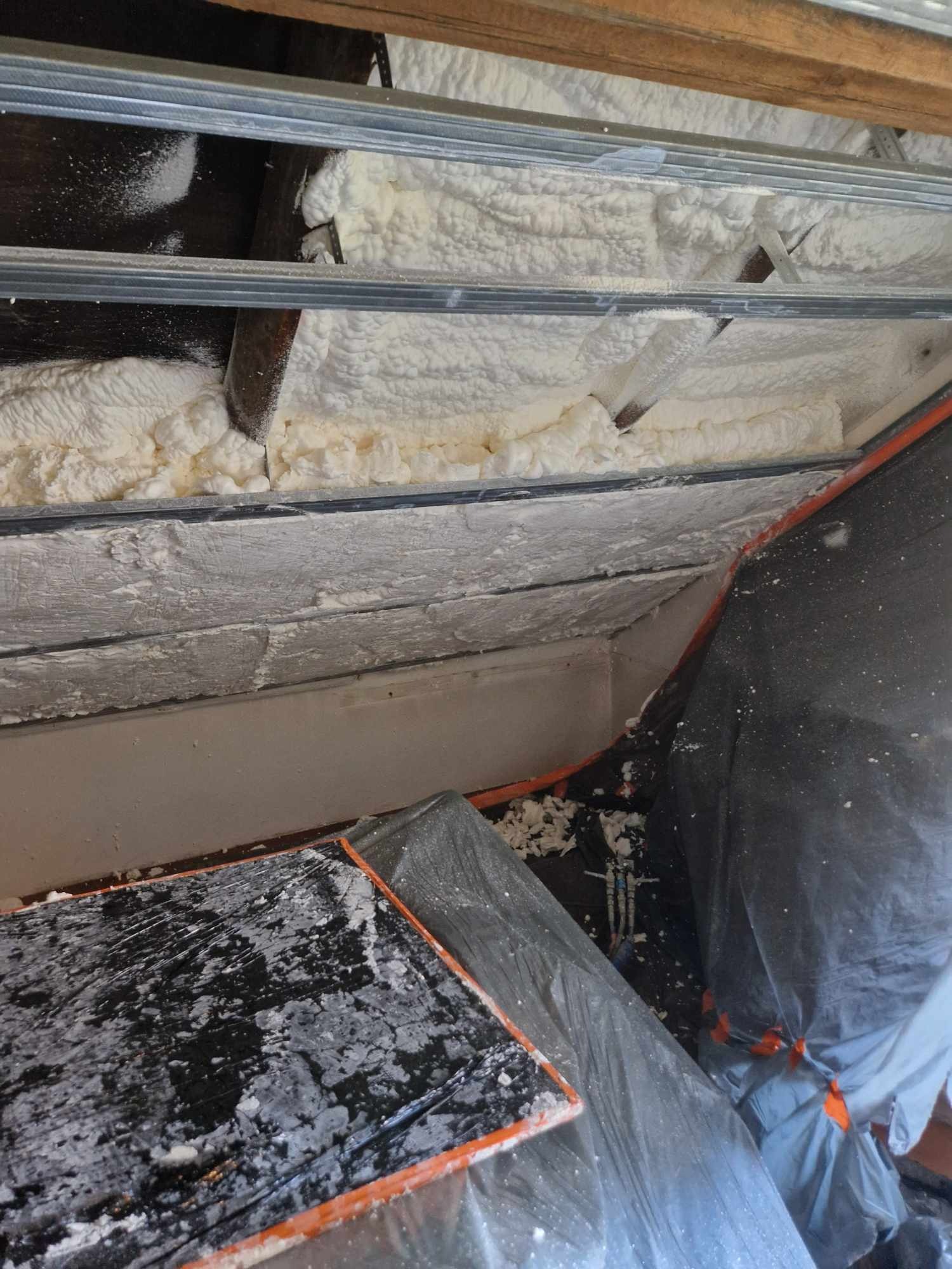 Poddasze z widoczną konstrukcją metalową i drewnianą, częściowo wypełnione białą pianą izolacyjną, zabezpieczone folią ochronną, widoczne kable.