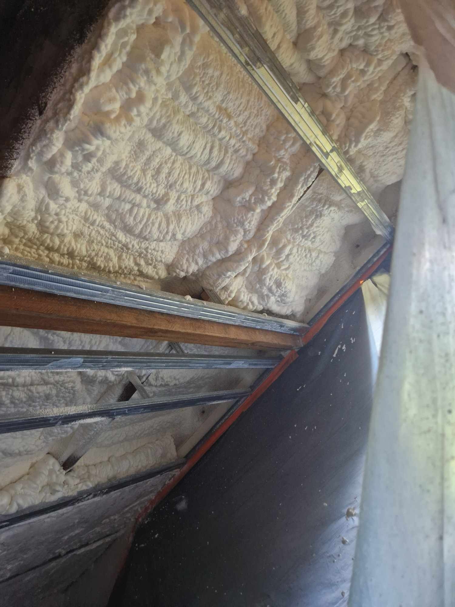 Poddasze z widoczną konstrukcją metalową i drewnianą, wypełnione jasną, spienioną izolacją natryskową o nieregularnej strukturze, częściowo osłonięte czarną folią i białą tkaniną.
