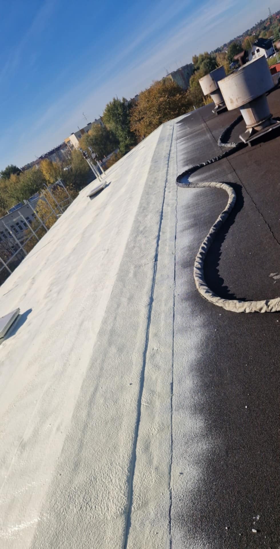 Płaski dach pokryty częściowo jasną pianą izolacyjną, z widocznymi kominami wentylacyjnymi i budynkami w tle, pod błękitnym niebem.