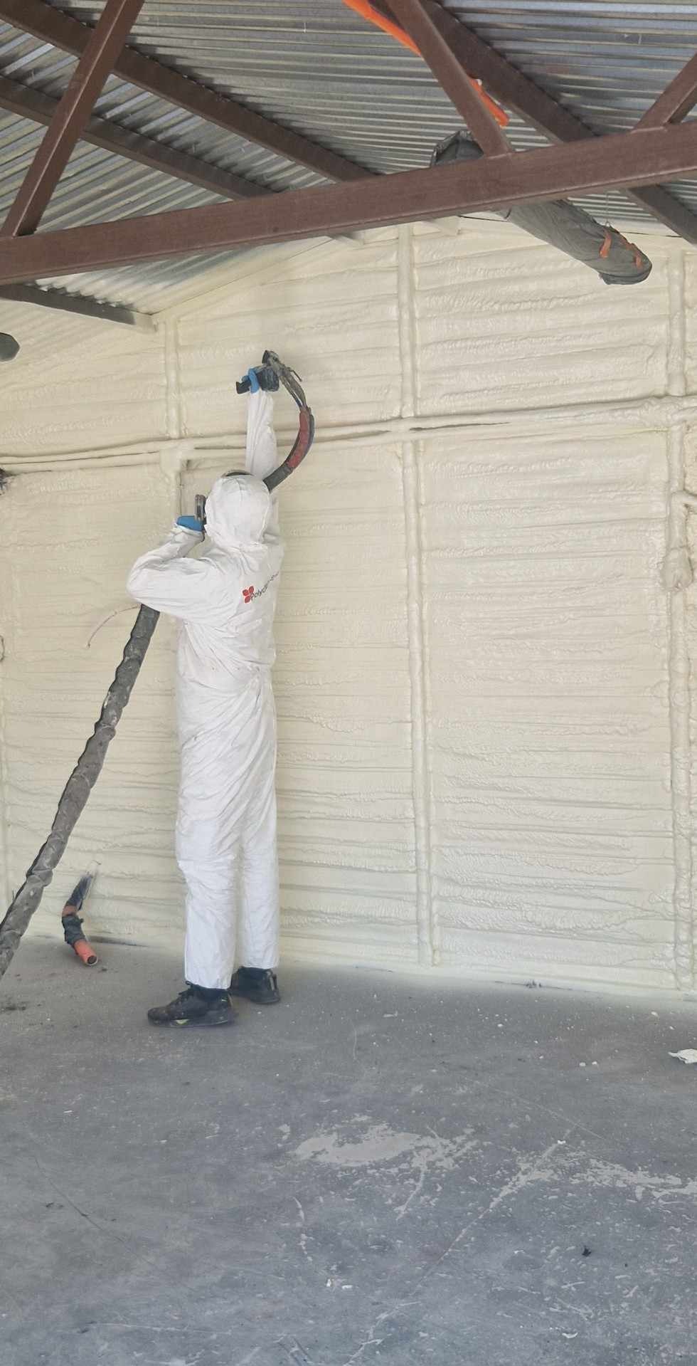 Pracownik w kombinezonie ochronnym natryskuje pianę izolacyjną na ścianę wewnątrz budynku z metalową konstrukcją dachu.