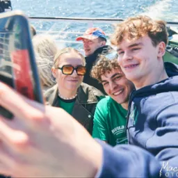 Grupa młodych ludzi robiąca selfie na promie, w tle widać wodę i innych pasażerów.