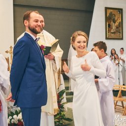 Maciej Ziętarski - Uśmiechnięta panna młoda w prostej białej sukni daje kciuk w górę podczas ceremonii ślubnej w kościele, obok stoi pan młody w niebieskim garniturze, w tle ministranci i chór.