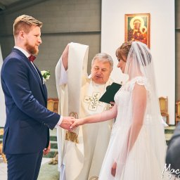 Maciej Ziętarski - Para młoda podczas ceremonii ślubnej w kościele, trzymają się za ręce, ksiądz w ornacie z krzyżem, w tle ikona i drewniane krzesła.