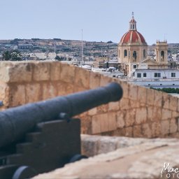 Maciej Ziętarski - Stara armata na kamiennym murze obronnym z widokiem na miasto z kościołem z czerwoną kopułą w tle.