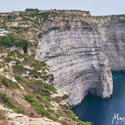 Maciej Ziętarski - Widok klifów z warstwowymi formacjami skalnymi opadającymi do ciemnoniebieskiego morza; biały samochód na drodze na szczycie klifu, roślinność na pierwszym planie.