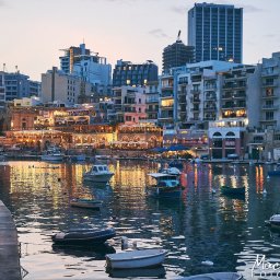 Maciej Ziętarski - Maltese port w wieczornym świetle: łodzie kołyszą się na wodzie odbijającej światła budynków i latarni. W tle zabudowa miejska z restauracjami i sklepami.