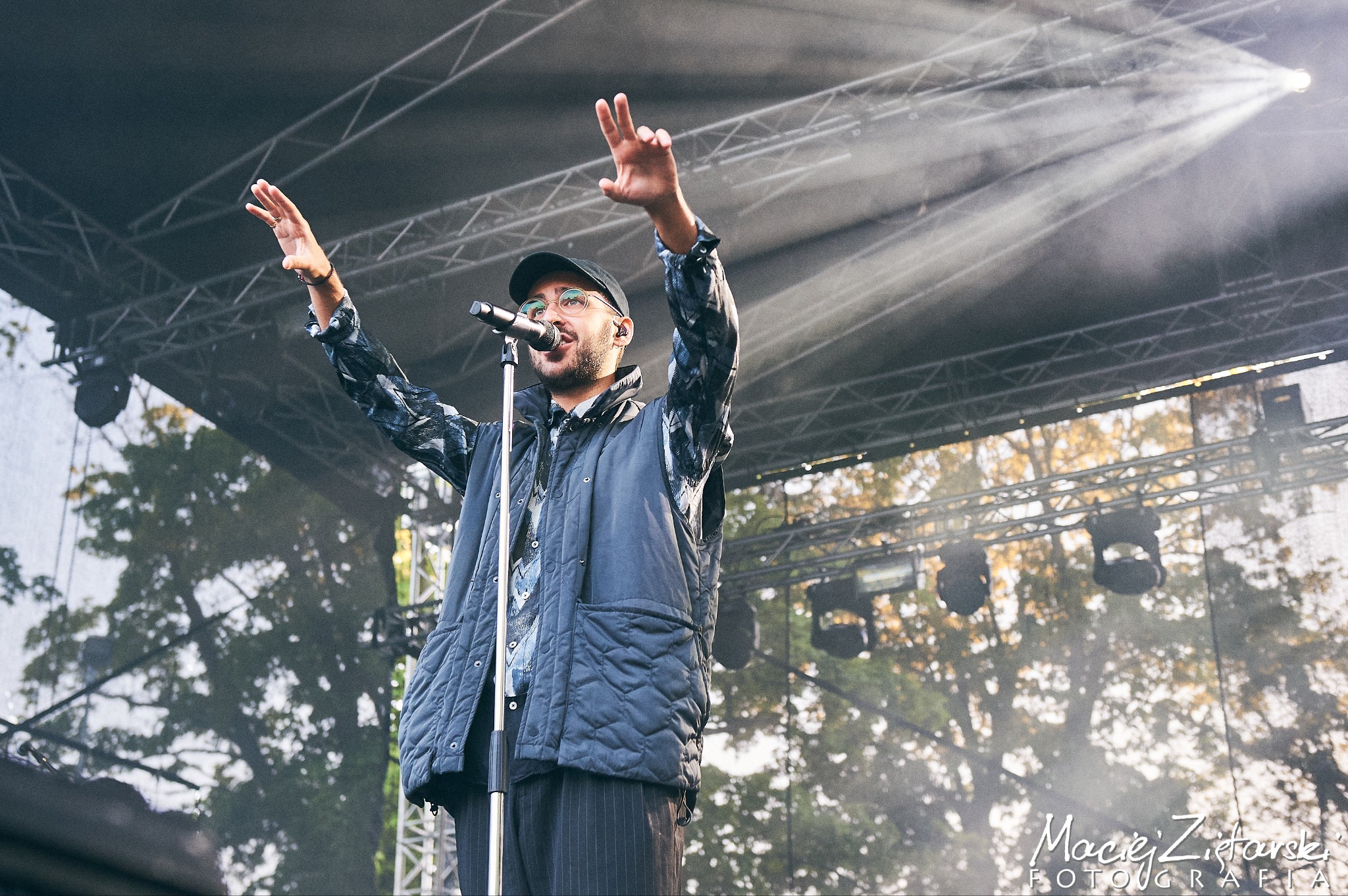 Wokalista z mikrofonem na scenie podczas koncertu plenerowego, oświetlony reflektorami, w tle drzewa.