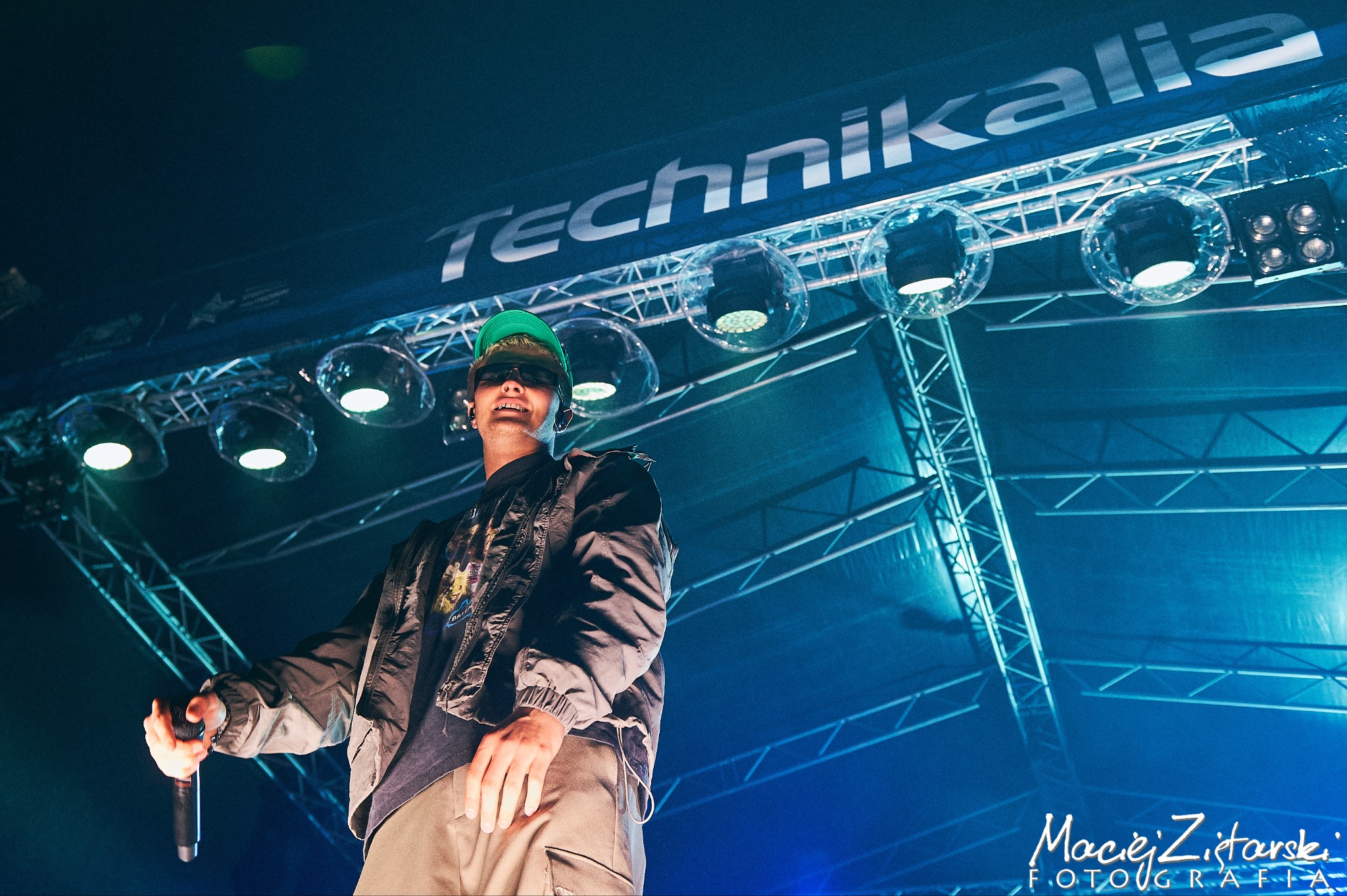 Artysta w zielonej czapce i okularach przeciwsłonecznych, trzymający mikrofon na scenie oświetlonej reflektorami i dekoracjami podczas występu.