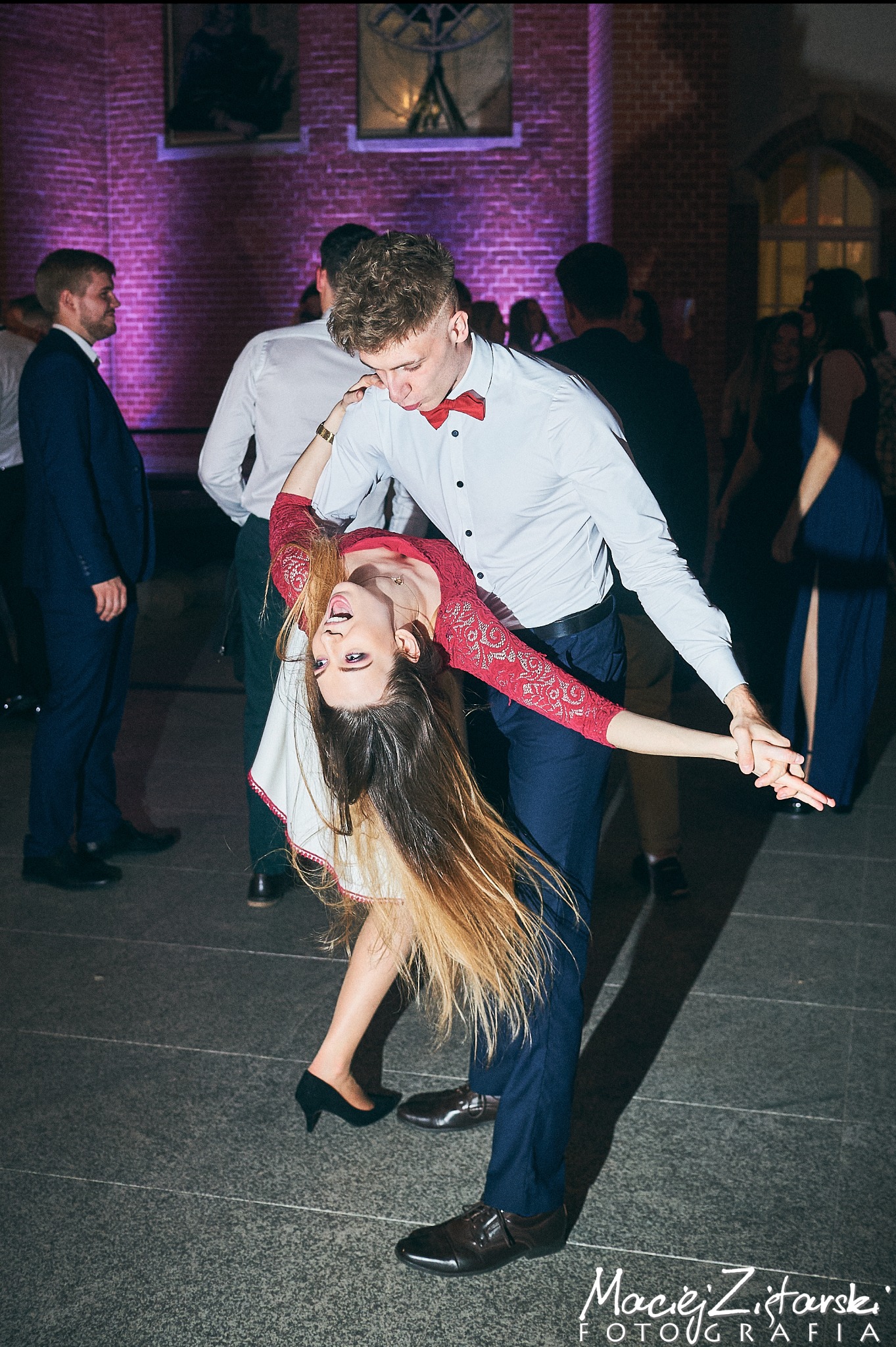 Para w dynamicznym ujęciu tanecznym na tle ceglanej ściany z fioletowym oświetleniem, ona w koronkowej sukience i szpilkach, on w białej koszuli i muszce, otoczeni innymi gośćmi na parkiecie.