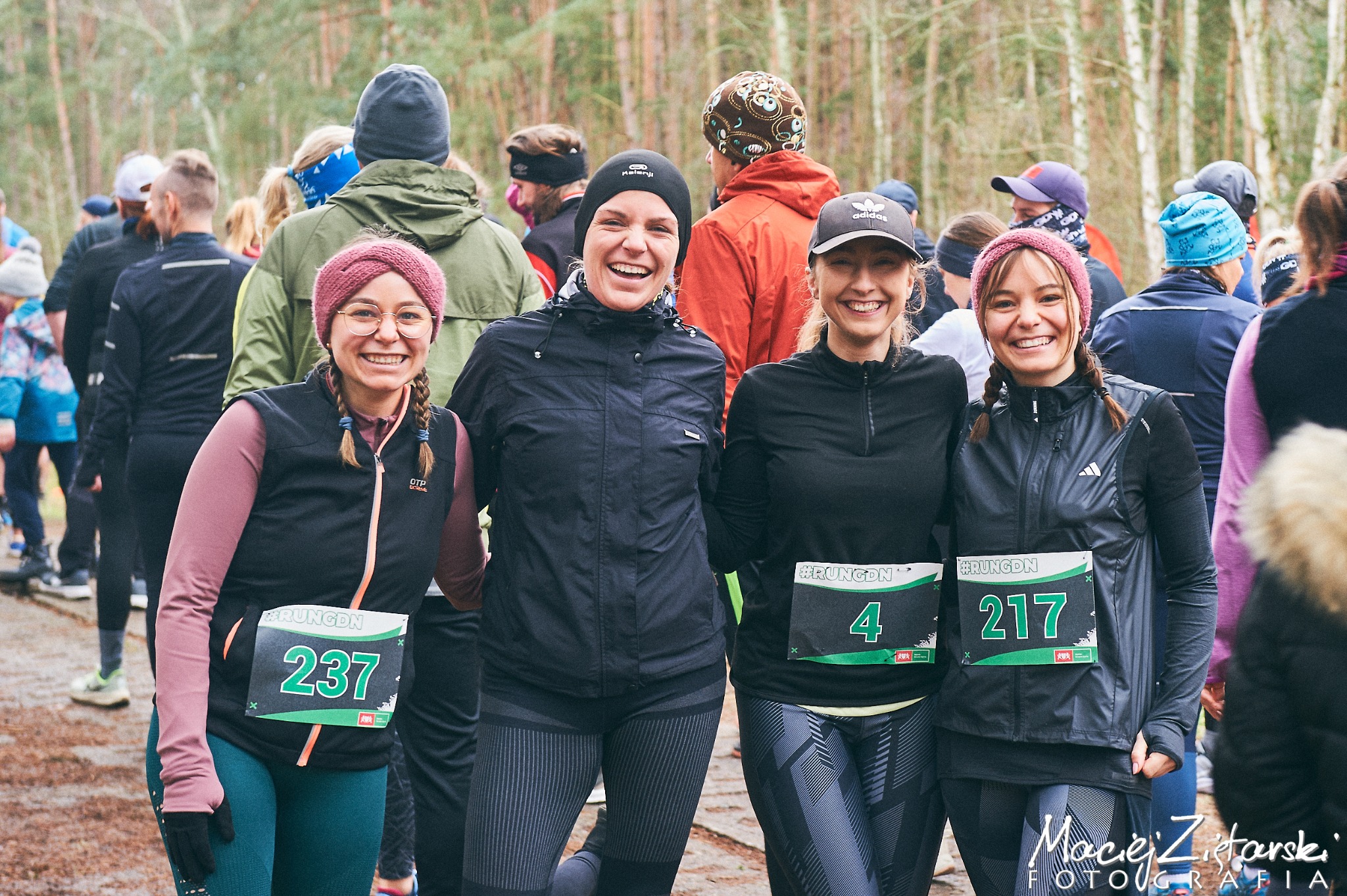 Grupa czterech uśmiechniętych kobiet w odzieży sportowej z numerami startowymi po biegu w lesie, widoczny tłum biegaczy w tle.