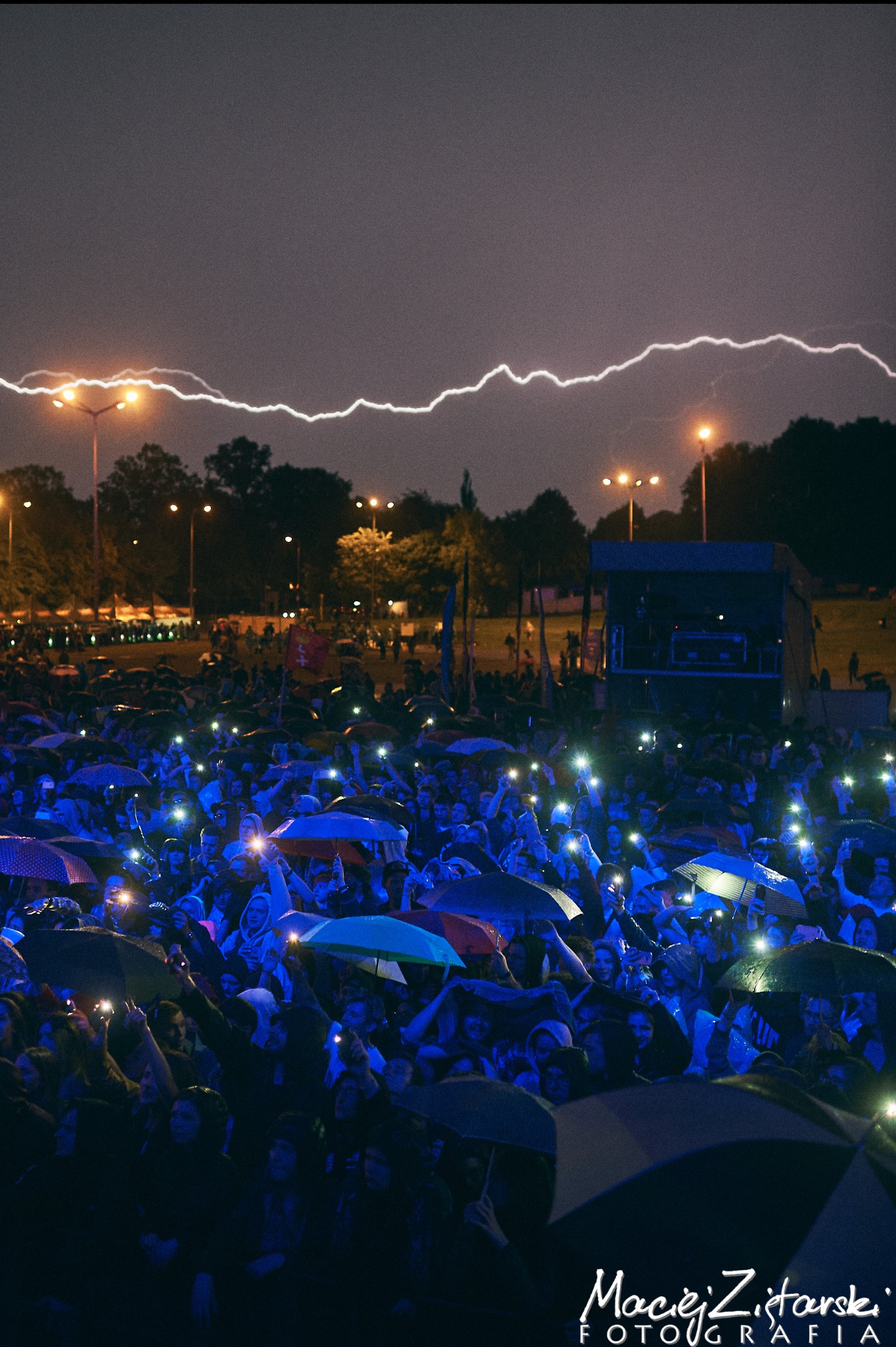 Tłum ludzi z parasolami i telefonami na koncercie plenerowym podczas burzy, z błyskawicą na nocnym niebie