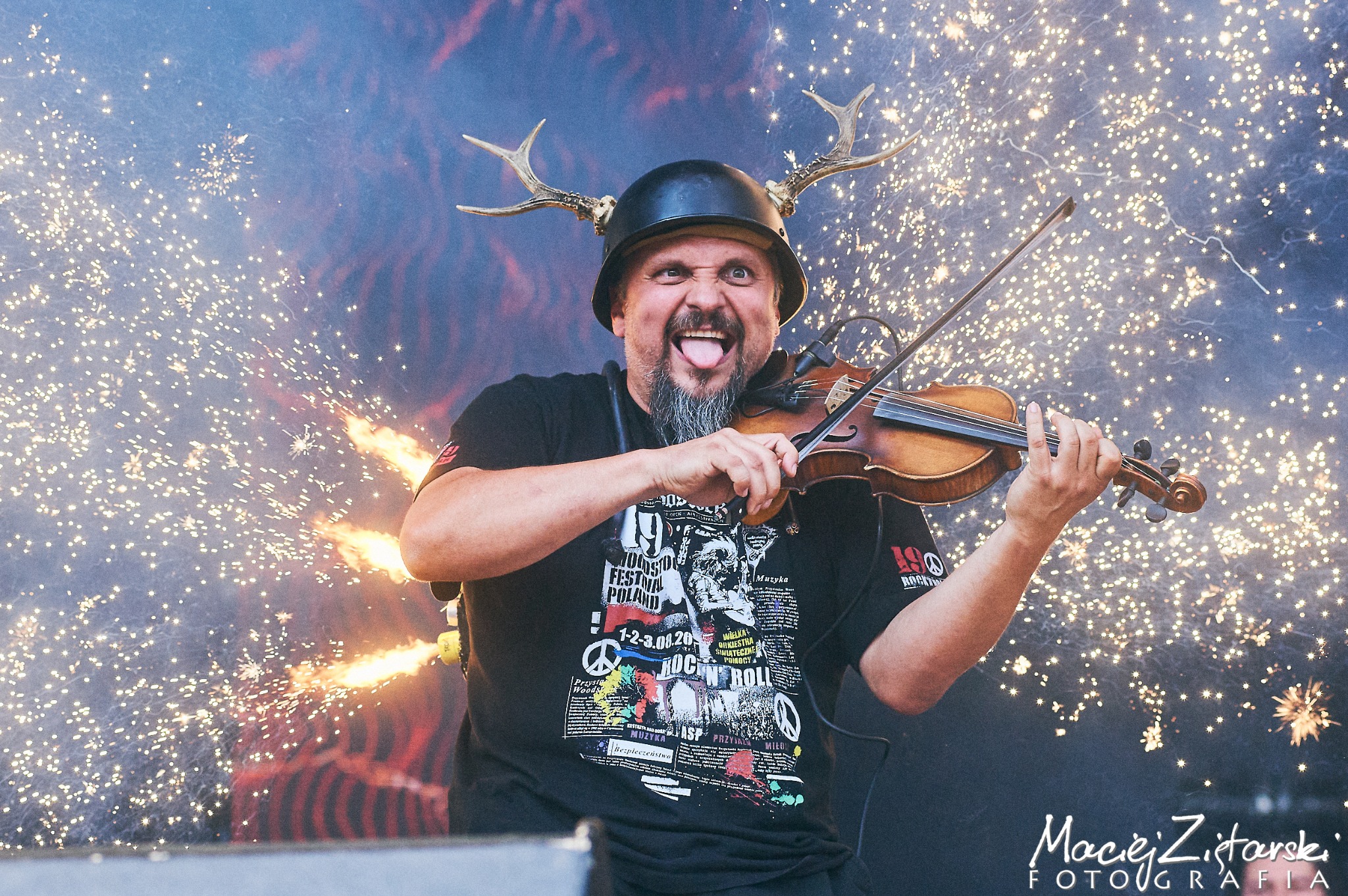 Ekscentryczny skrzypek z hełmem i rogami jelenia na scenie, wystawia język, w tle efekty pirotechniczne i iskry.