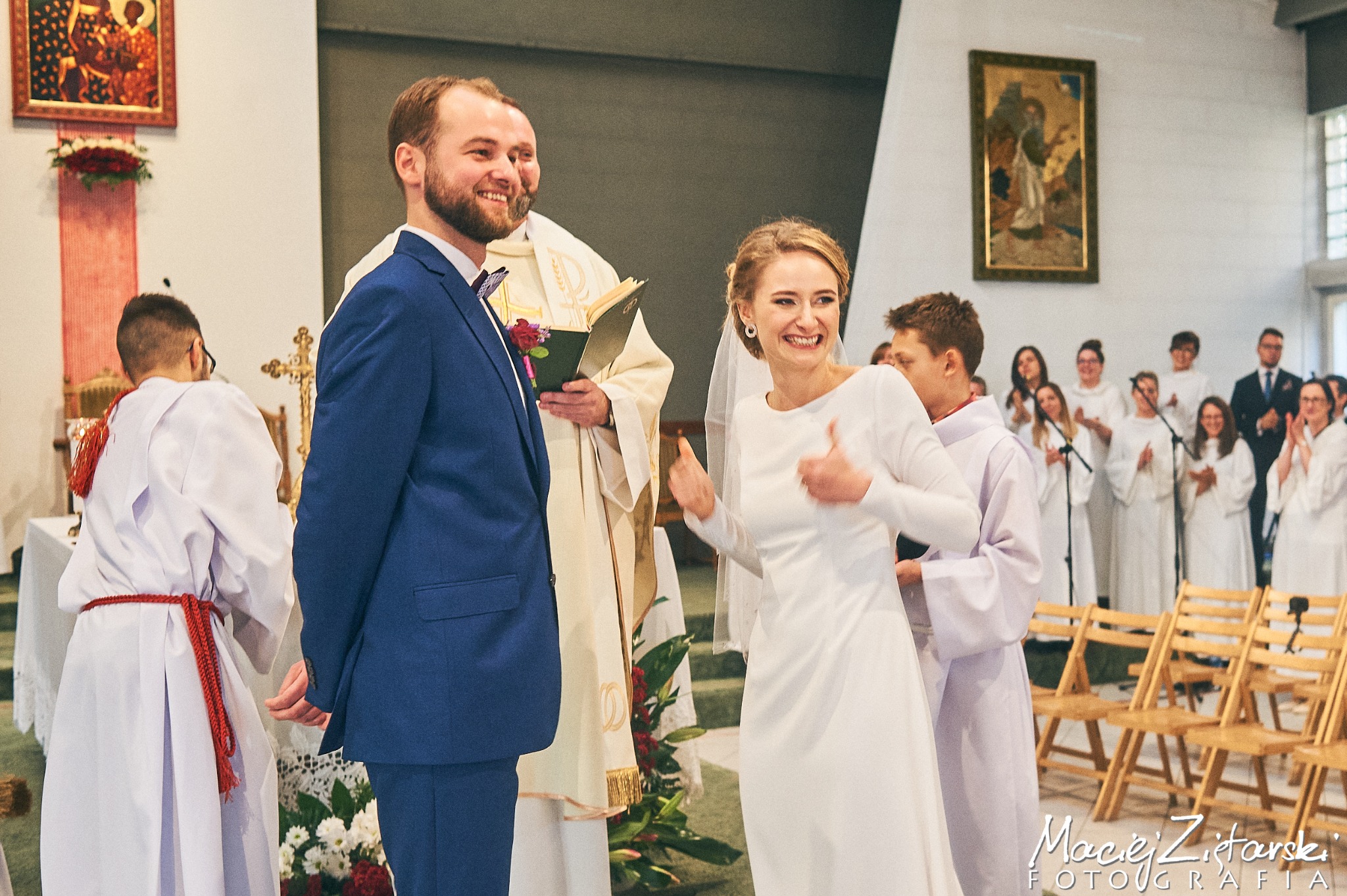 Uśmiechnięta panna młoda w prostej białej sukni daje kciuk w górę podczas ceremonii ślubnej w kościele, obok stoi pan młody w niebieskim garniturze, w tle ministranci i chór.