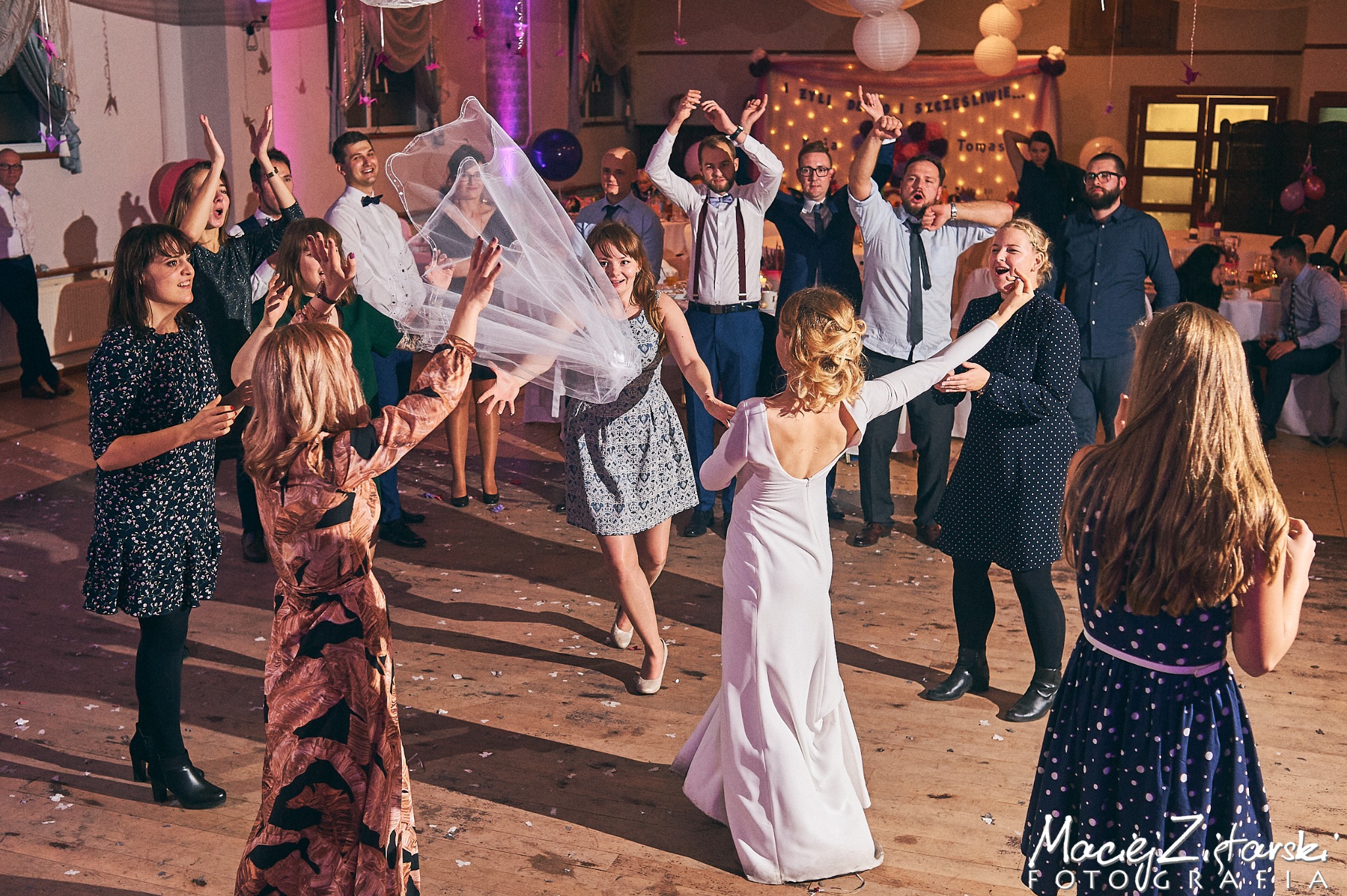 Panna młoda w białej sukni rzuca welon w tłum gości weselnych na parkiecie, radosne uniesienie, dekoracje balonowe i świetlne w tle.