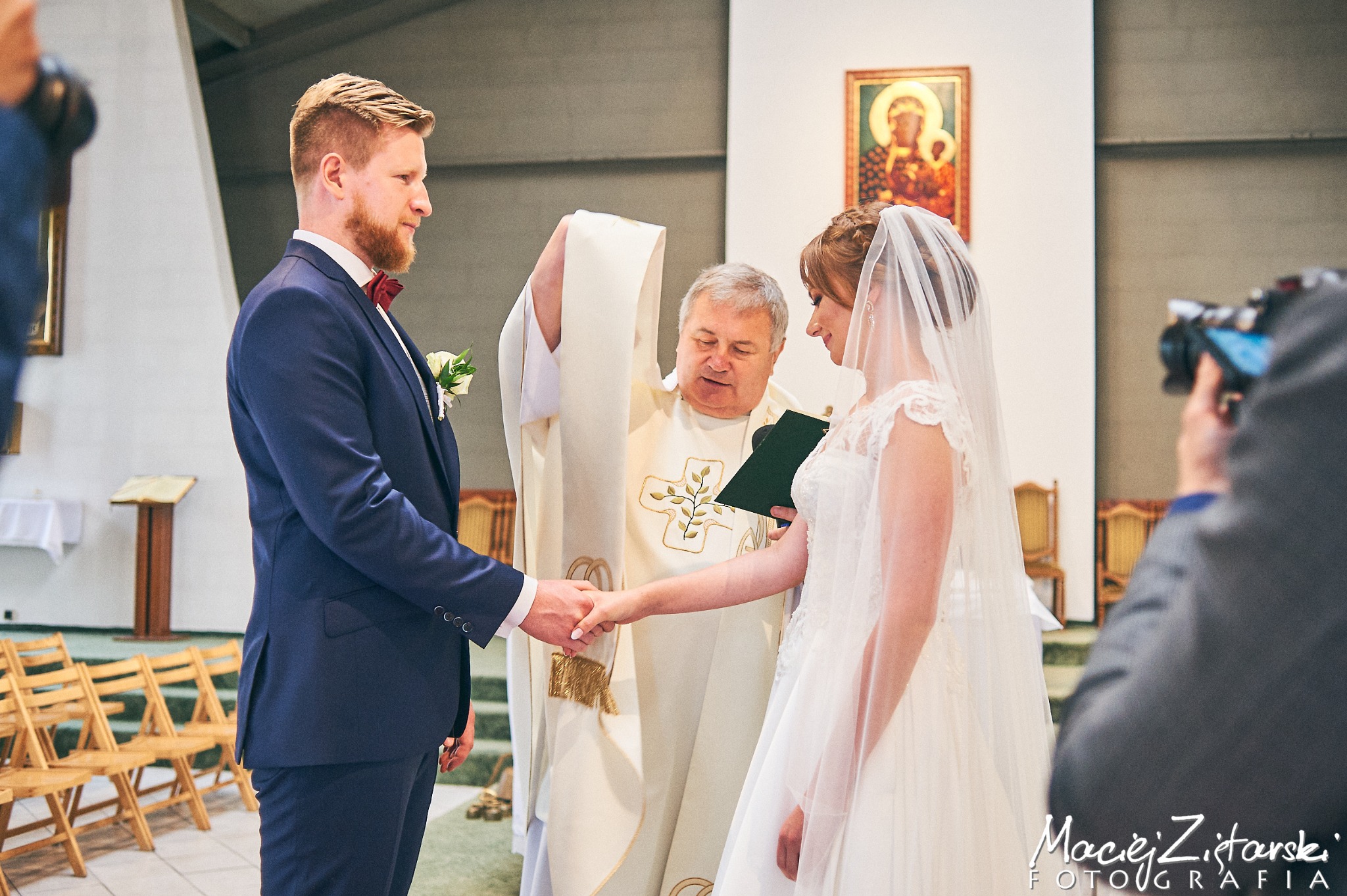 Para młoda podczas ceremonii ślubnej w kościele, trzymają się za ręce, ksiądz w ornacie z krzyżem, w tle ikona i drewniane krzesła.