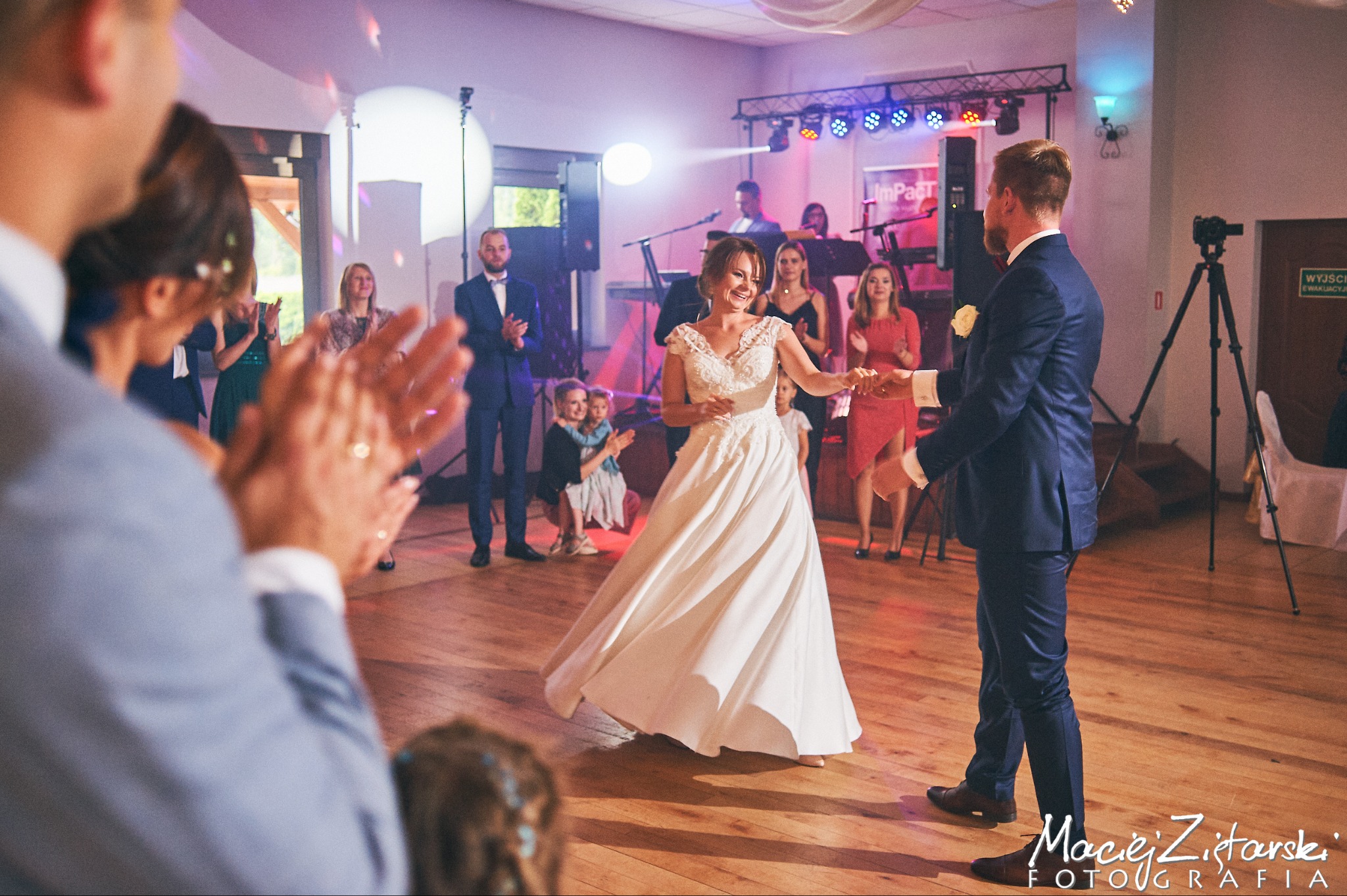 Panna młoda w białej sukni tanecznej, uśmiechnięta, trzyma kartkę podczas pierwszego tańca z panem młodym w granatowym garniturze, goście klaszczą w tle, widoczna scena z zespołem i oświetleniem.