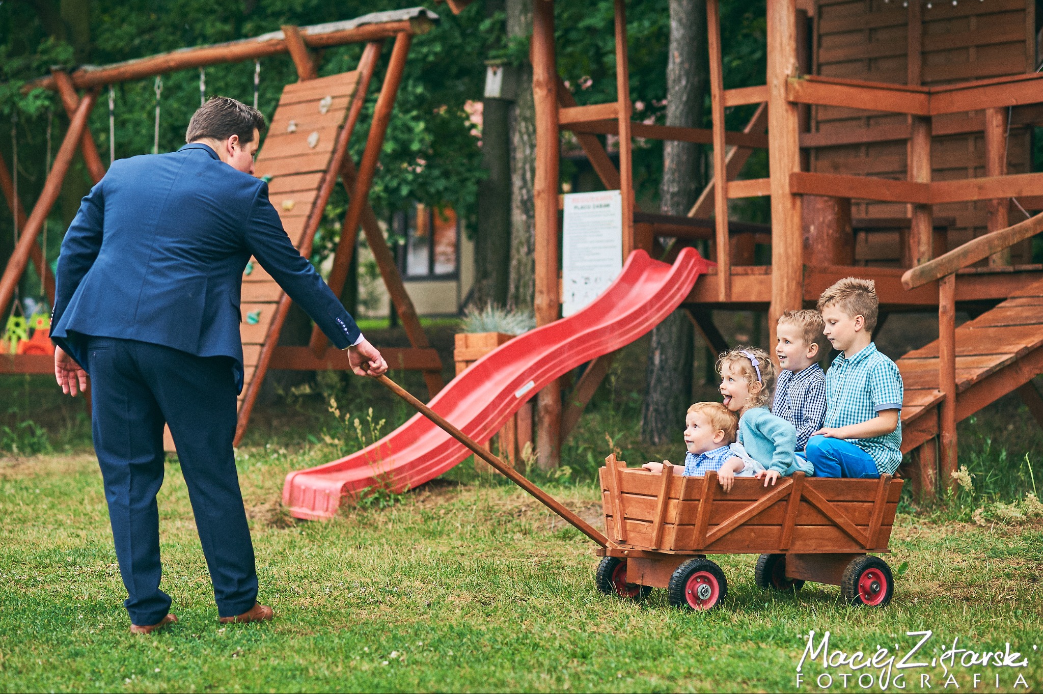 Mężczyzna w garniturze ciągnie drewniany wózek z czwórką dzieci na tle placu zabaw z drewnianą konstrukcją i czerwoną zjeżdżalnią.