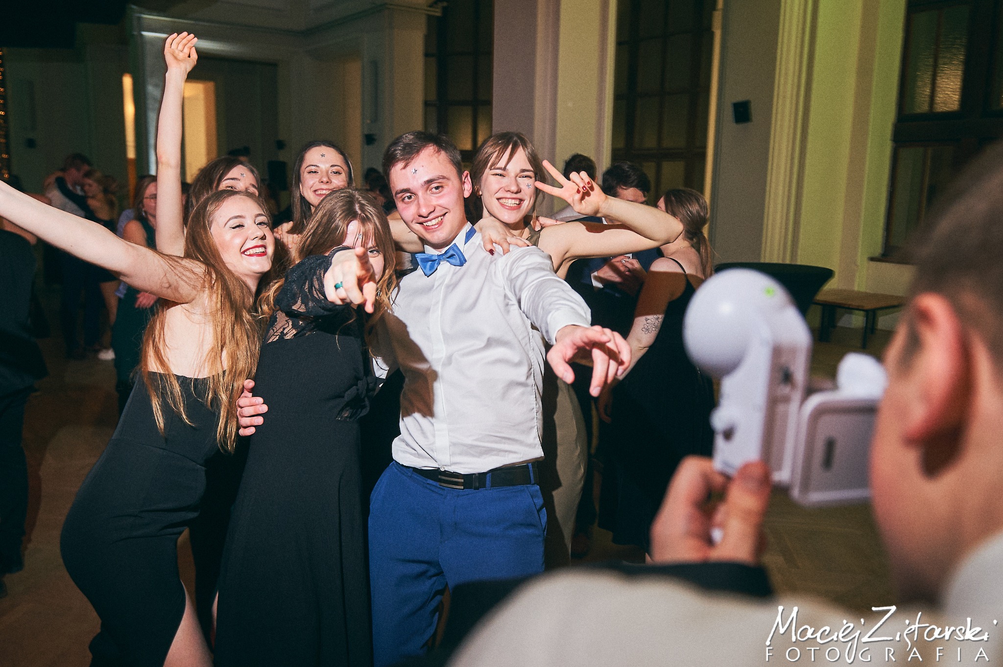 Grupa roześmianych młodych ludzi pozuje do zdjęcia na tle sali balowej, z widocznym fotografem trzymającym aparat w dolnym rogu kadru.