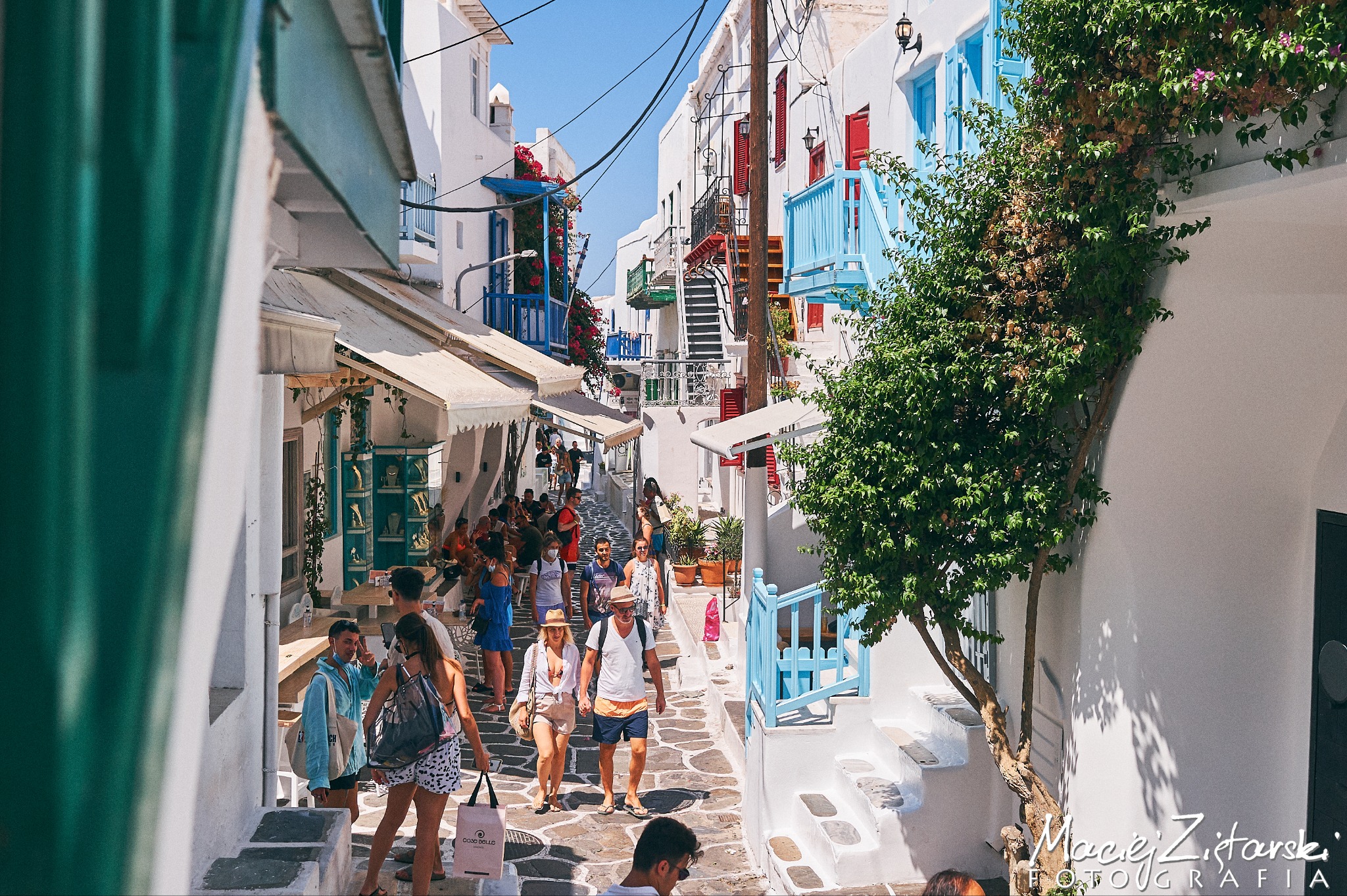 Ulica w greckim stylu z białymi budynkami, niebieskimi akcentami i przechodzącymi turystami w słoneczny dzień.