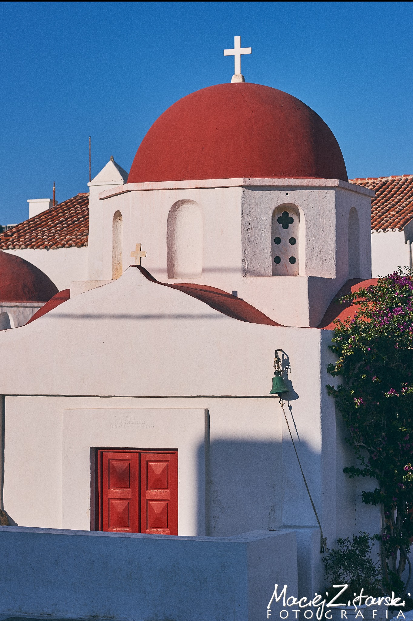Biały kościół z czerwoną kopułą i krzyżem na tle błękitnego nieba, widoczne czerwone drzwi i dzwon.