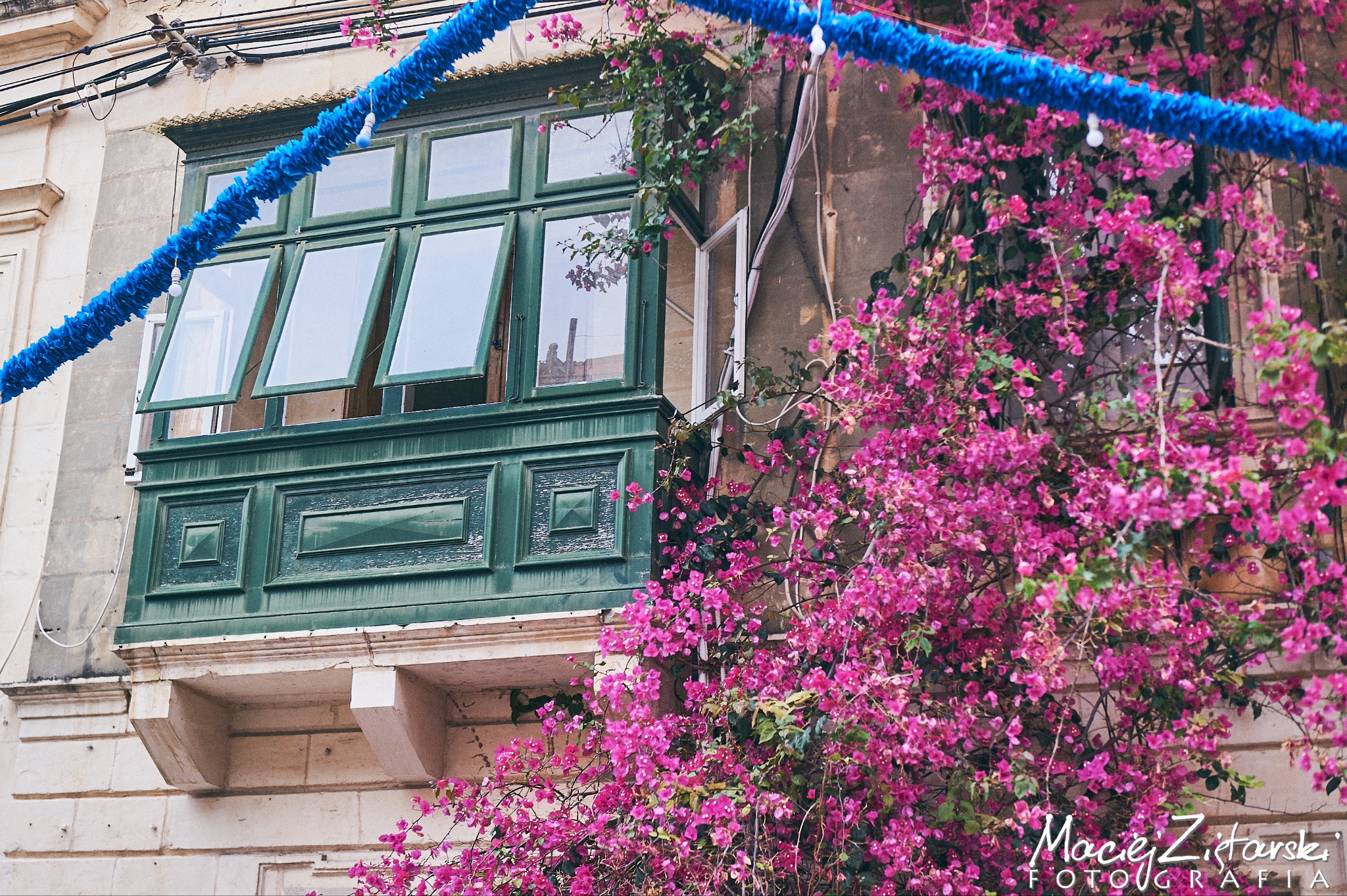 Zielony balkon z otwartymi oknami, porośnięty kwitnącą bugenwillą, udekorowany niebieską girlandą. Widoczne detale architektoniczne budynku.