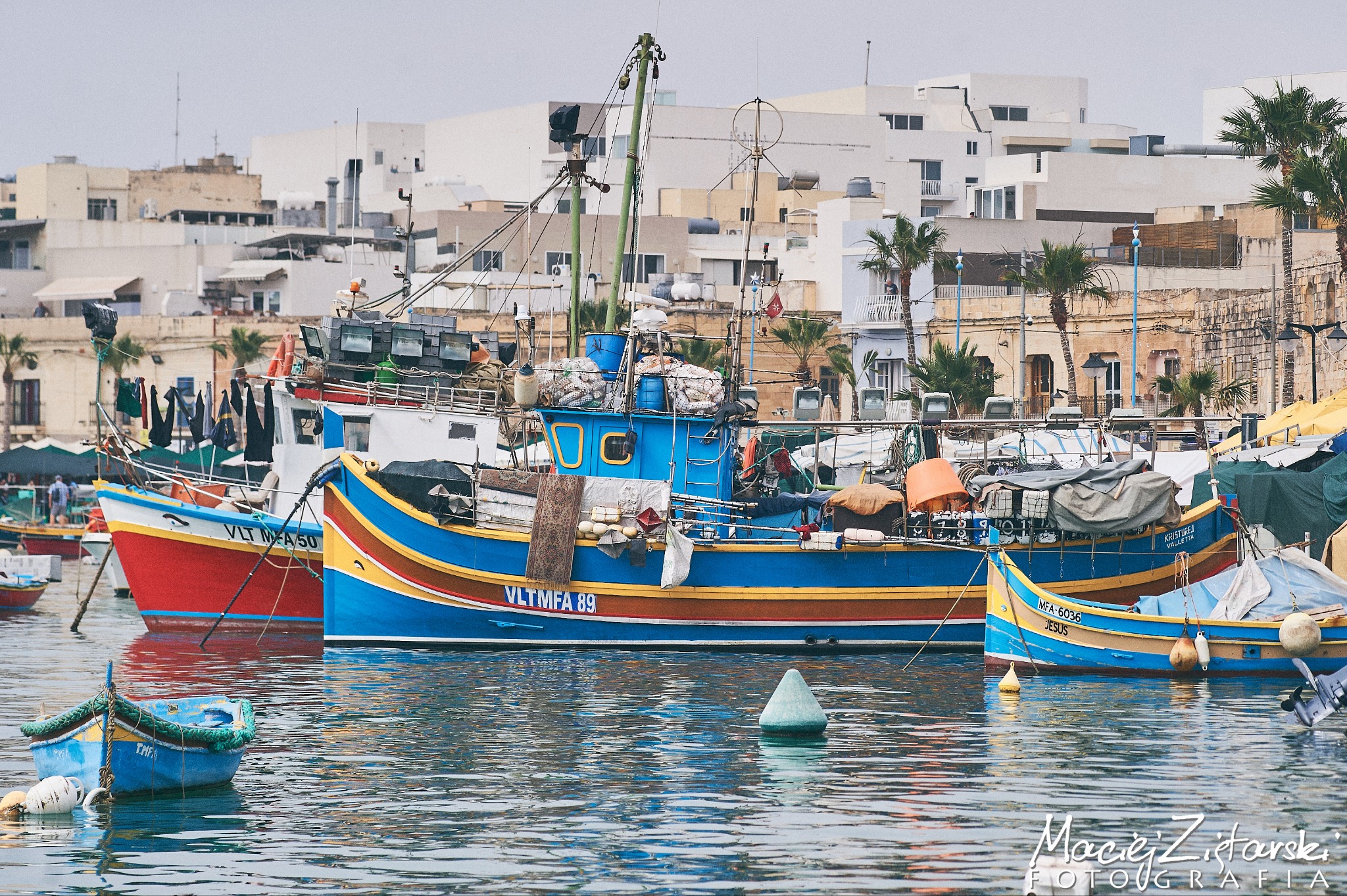 Kolorowe łodzie rybackie z nazwami VLT MFA 89, VLT MA 50 i JESUS zacumowane w porcie na tle zabudowy miejskiej i palm.