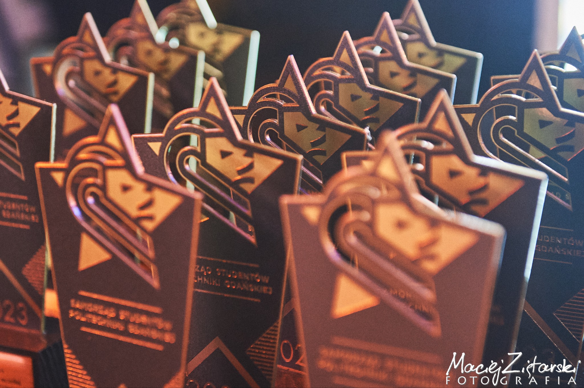 Rząd metalowych statuetek z motywami geometrycznymi i wizerunkiem twarzy, ustawionych blisko siebie, z widocznym logo fotografa w prawym dolnym rogu.
