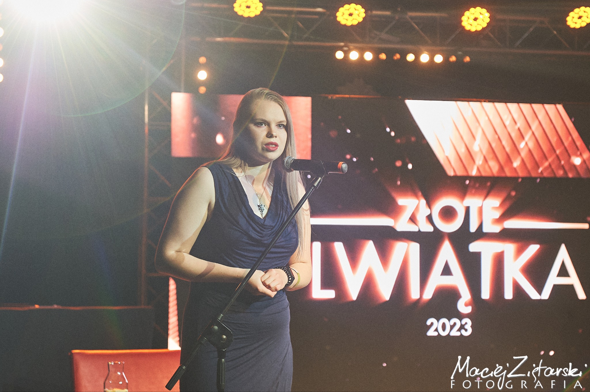 Kobieta przemawia do mikrofonu na tle podświetlonego napisu 'Złote Lwiątka 2023' na scenie eventowej z reflektorami.