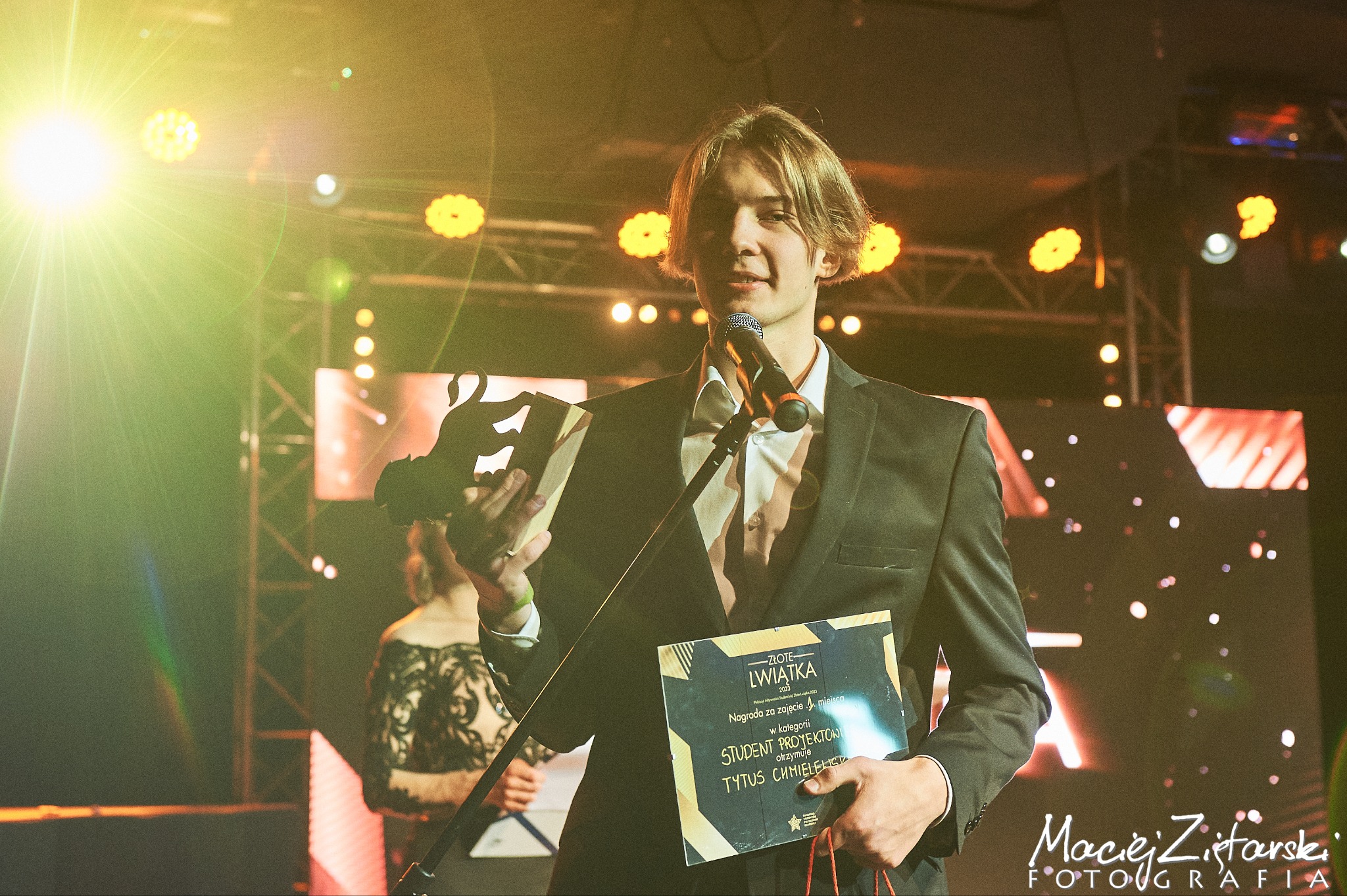 Młody mężczyzna w garniturze odbiera nagrodę 'Złote Lwiątka' za zajęcie 1 miejsca w kategorii Student Projektów, trzymając statuetkę i dyplom na scenie z oświetleniem eventowym.