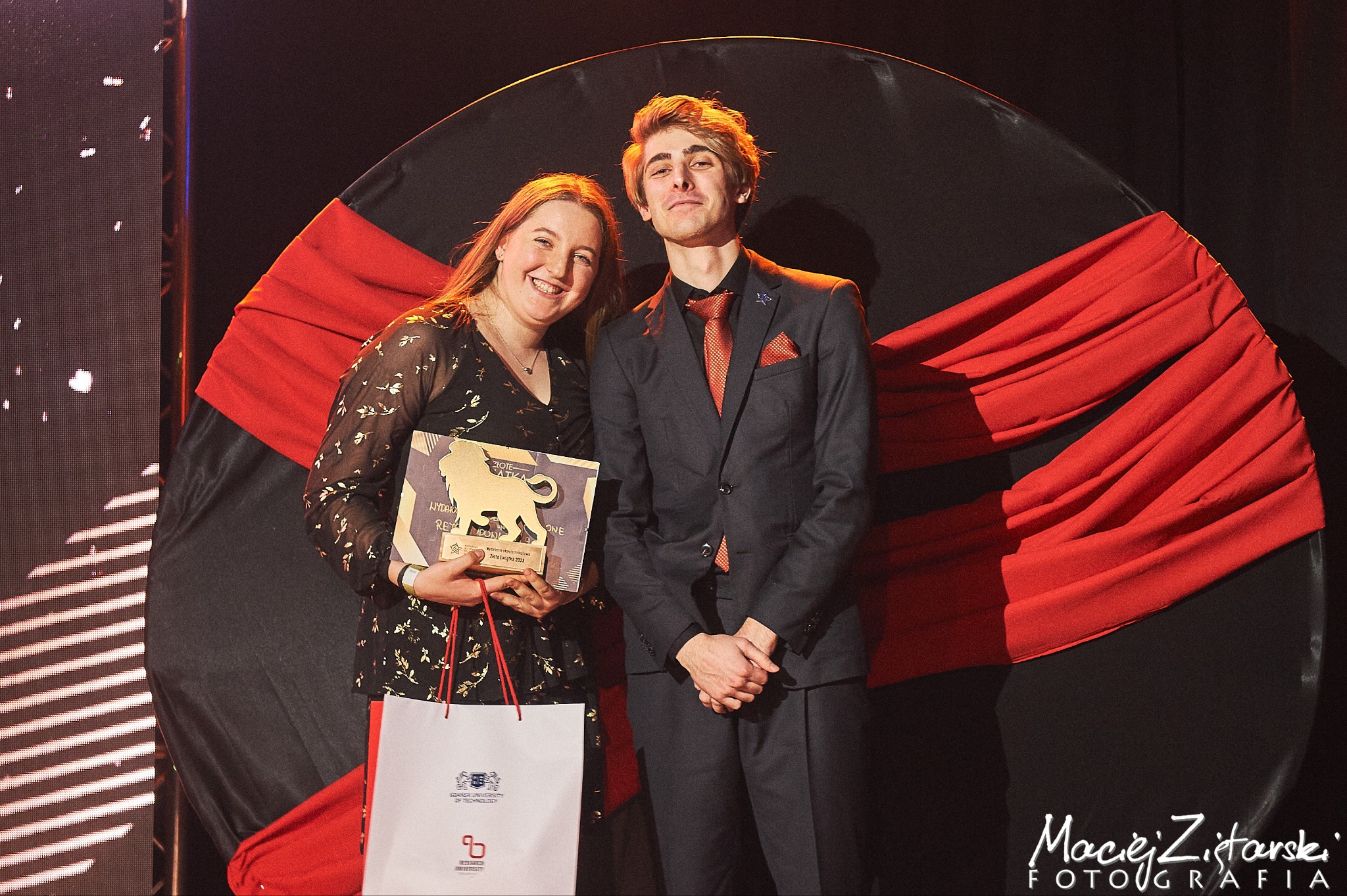 Dwoje młodych ludzi pozuje z nagrodą 'Złote Lwiątka' na tle sceny z czarnym kołem i czerwonym materiałem.