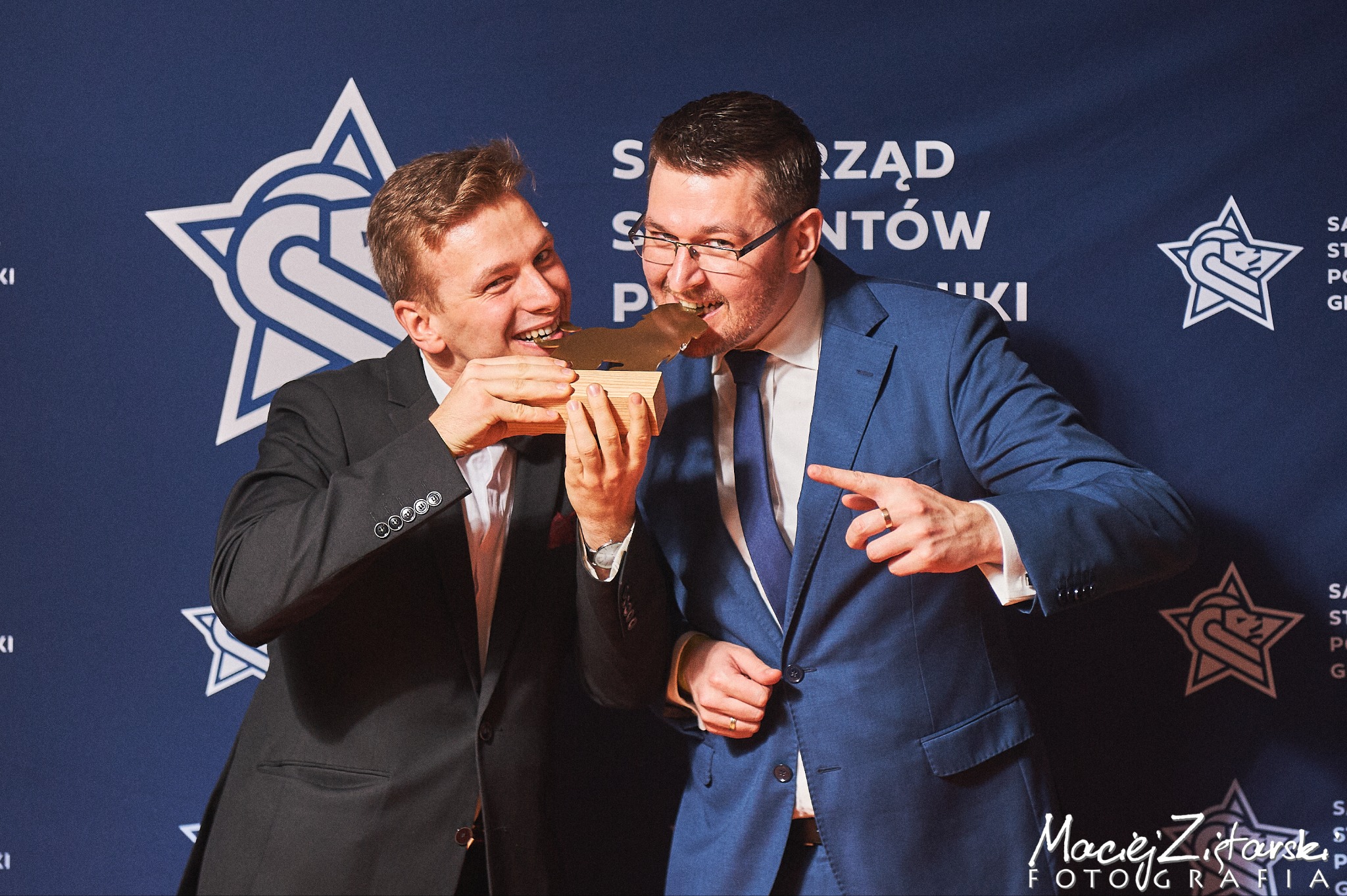 Dwóch mężczyzn w garniturach gryzie z uśmiechem złotą statuetkę na tle ściany z logo wydarzenia.