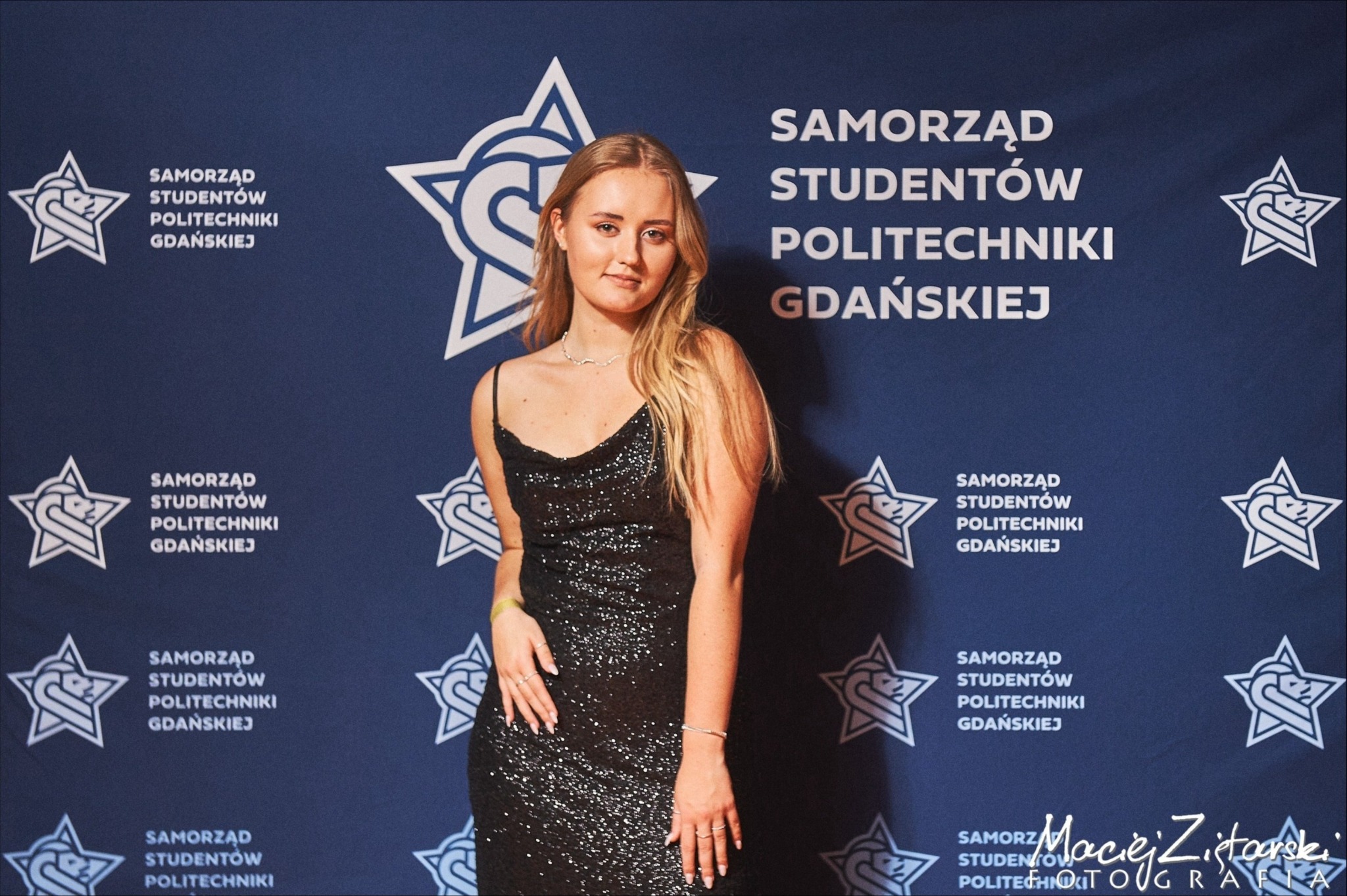 Portret kobiety w czarnej, błyszczącej sukience na tle ściany z logo Samorządu Studentów Politechniki Gdańskiej.