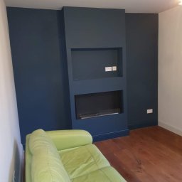 Prywatny wykonawca - Pomieszczenie po malowaniu ścian na ciemny granat z wnęką na telewizor i kominek elektryczny, drewniana podłoga i jasna kanapa.